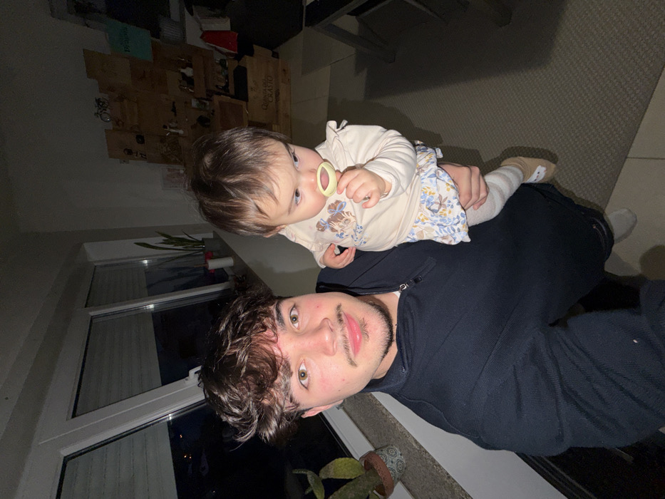 An indoor scene in Vila Real, Portugal, depicts a young adult male holding an infant at night. The male, positioned at the left, has dark curly hair, a mustache, and stubble, wearing a dark blue half-zip pullover, and is taking a selfie. He supports the infant, who is dressed in a light-colored patterned long-sleeve top, patterned bottoms, striped tights, and light-colored slippers with animal details. The infant holds a yellow-green pacifier or teether in its mouth and looks slightly to the lower right. Behind them, to the left, a large window with white frames shows the dark exterior, with a potted plant on the sill. To the right, a multi-tiered wooden display unit, appearing to be constructed from crates, holds bottles and various decorative objects, with the partial text "QUINTA DO CRAS" visible on one crate. The floor is covered with a light-colored patterned carpet or rug.