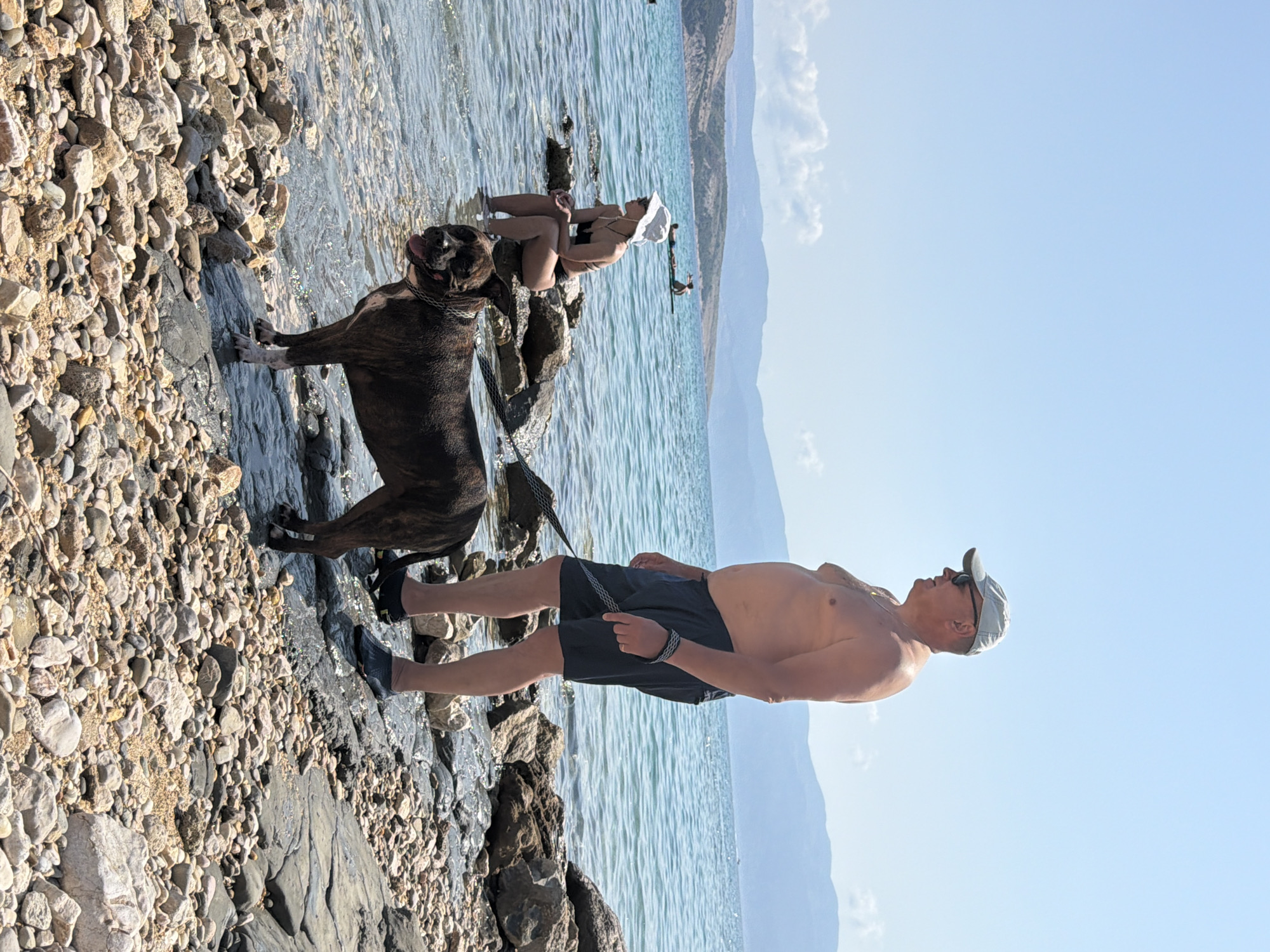 A full shot captures a man and a dog on a rocky shore next to a body of water under a clear blue sky. The man, with a tan complexion, is shirtless and wearing a light-colored baseball cap and dark shorts. He stands on the right, looking to the left. His right hand is holding a leash connected to a brindle-colored dog standing in the foreground. The dog faces forward, tongue out, appearing happy.
To the left of the dog, a woman sits on rocks in the shallow water. She wears a black bikini and a white sun hat. Her legs are submerged in the water, and she is looking out towards the sea. In the background, two people are visible on paddleboards in the blue water. Further back, mountains rise against the horizon. The shore is covered in a mix of small and large rocks. The water is calm and clear, with light reflecting off its surface. The scene appears to be a beach in Općina Baška, Croatia.