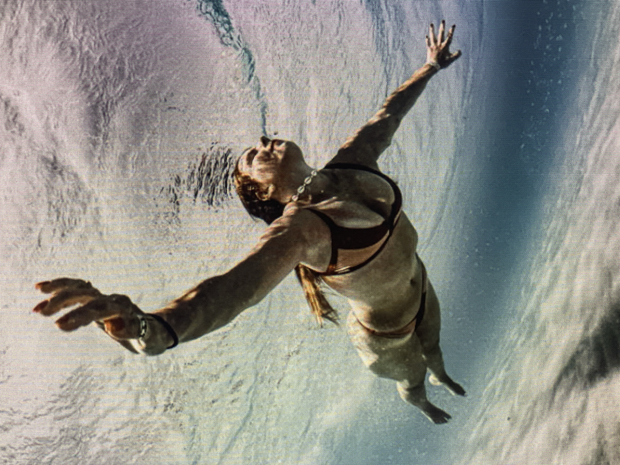 An underwater shot captures a woman with red hair swimming towards the surface. She is wearing a dark bikini and appears to be exhaling as bubbles rise from her mouth. Her right arm is extended upwards, with her hand open. The water is clear and illuminated by sunlight filtering from above, creating patterns of light and shadow on the sandy bottom.