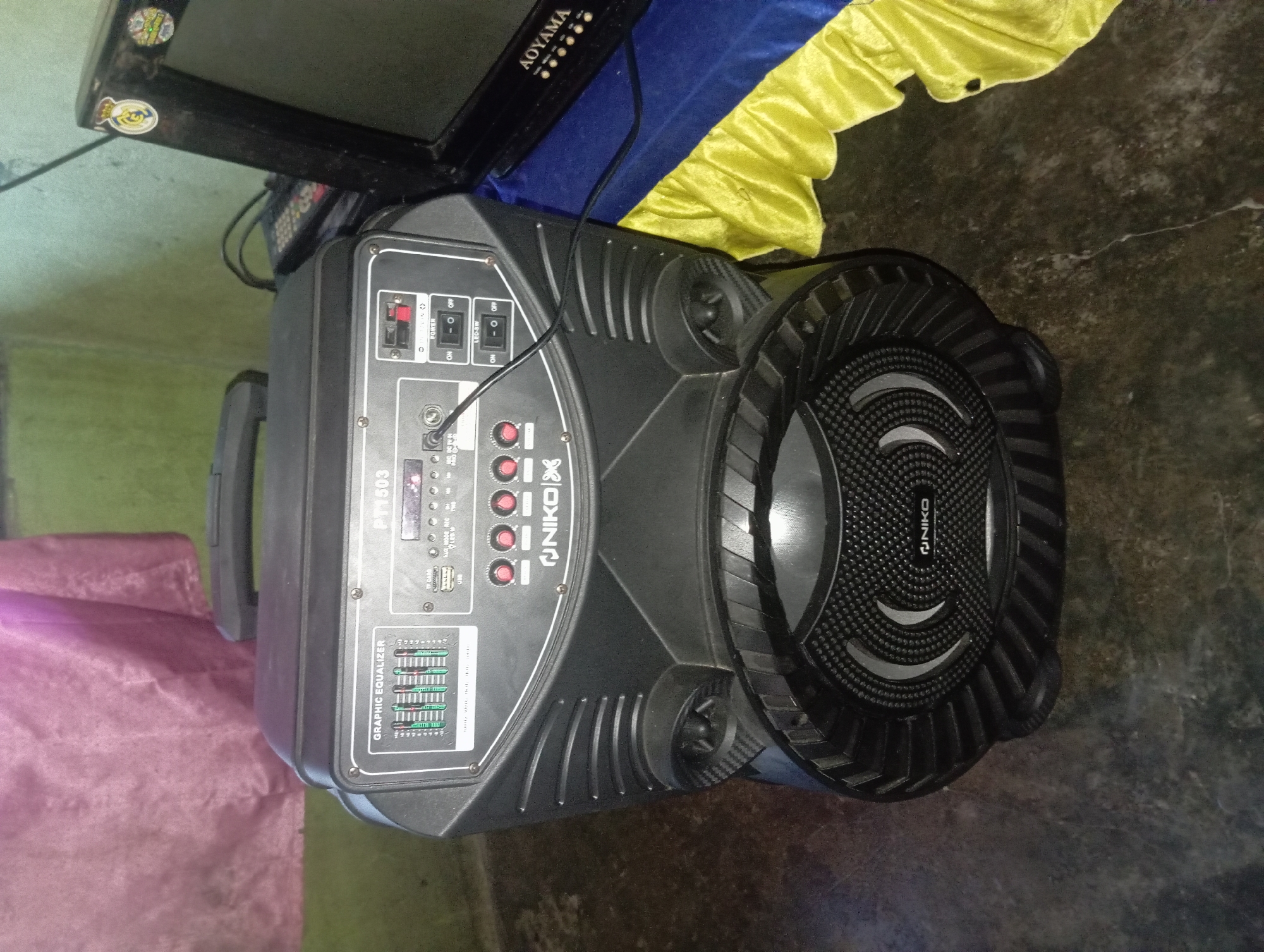 This is a close-up, eye-level, indoor shot of a black portable speaker in what appears to be a room in Rejosari, Indonesia. The speaker is large, with a handle on top and a prominent round speaker grill at the bottom. The top panel of the speaker has various controls and inputs, including a graphic equalizer display, buttons, knobs, and ports for USB, AUX, and DC power. The model number "PT1503" is visible on the speaker. To the right of the speaker, a vintage television set with an "AOYAMA" logo is partially visible. The television is adorned with two round stickers, one depicting a royal crown with "R" inside, and another with a shield insignia. The background consists of a textured floor and a wall, with a hint of a draped pink fabric on the left and a blue and yellow cloth draped on the right, likely covering the base of the television. The lighting is dim, suggesting it might be evening or the room is not brightly lit. There are no people or active interactions depicted in the scene.