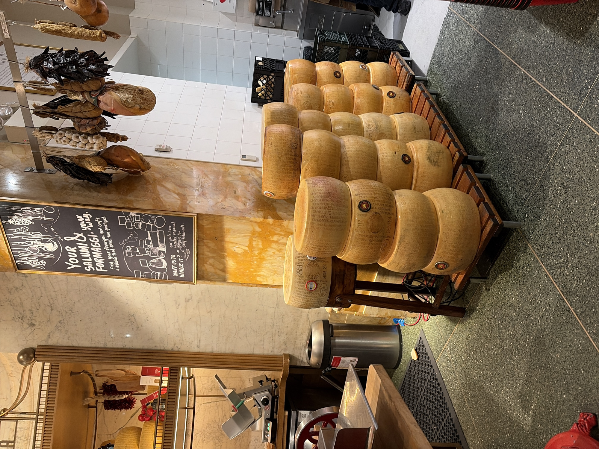 This image depicts the interior of an Italian deli or specialty food store, likely in New York City, United States of America. In the foreground, several large wheels of cheese, appearing to be Parmesan or a similar hard cheese, are stacked on wooden shelves. To the left, a professional meat slicer is visible on a stainless steel counter. Behind the slicer, a cylindrical metal trash can stands next to a wooden shelving unit displaying some packaged food items and what appear to be dried chili peppers.
Hanging from a metal rack above the cheese are various cured meats and sausages, typical of an Italian salumi selection. A chalkboard sign hangs on the wall to the left, with handwritten text in English advertising "Your Salumi & Formaggi" and offering to help customers find their favorites. The walls are adorned with marble-like tiles and white subway tiles in the background, suggesting a clean and professional food service environment. The floor is a speckled green terrazzo. The overall impression is a well-stocked, authentic Italian food establishment.