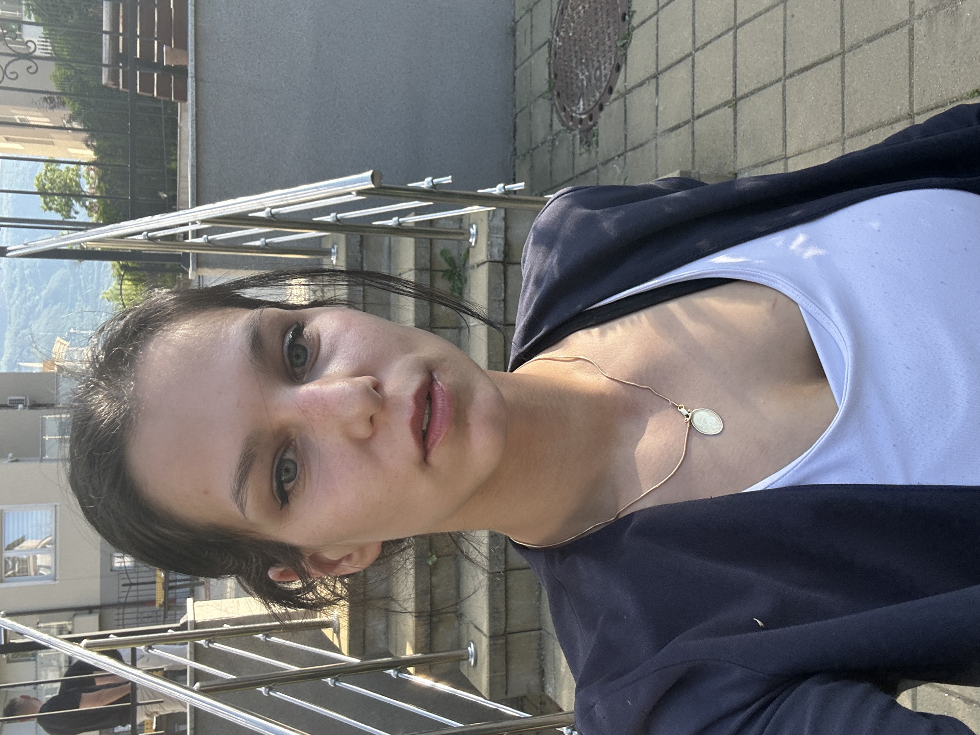 A medium close-up photograph depicts a young woman, likely in her early twenties, with dark hair and fair skin. She is wearing a white top under a dark-colored jacket. A delicate gold necklace with a circular pendant adorns her neck. She is facing the camera with a neutral expression.
The background reveals an outdoor setting with modern architecture. A metallic railing suggests she might be on a staircase or balcony. Stone paving is visible in the background, along with glimpses of buildings and foliage. Based on the architectural style and the presence of mountains in the distance, the location is speculated to be within the city of Sofia, Bulgaria. A manhole cover is visible on the stone paving.