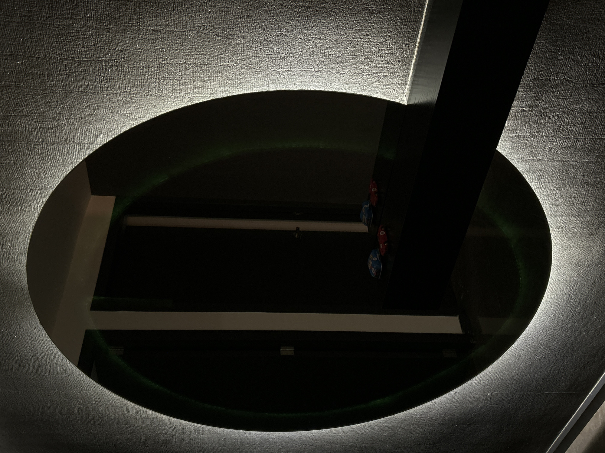 A low-angle, close-up photograph shows an oval-shaped mirror mounted on a textured surface that appears to be a ceiling. The mirror reflects a dark interior space with horizontal linear elements, likely architectural details such as beams or shelves. Several small, possibly decorative items (red and blue) are visible in the reflection toward the upper-right area of the oval shape. A dark, vertical rectangular shape crosses the upper-right portion of the mirror, partially obscuring the reflected interior space. The periphery of the mirror is illuminated with a soft, white light, and a faint green glow is seen along the inner edge of the oval. The textured ceiling surface is a grayish-white. The photograph was likely taken indoors. The location of the image is San Bernardino Tlaxcalancingo, Mexico.