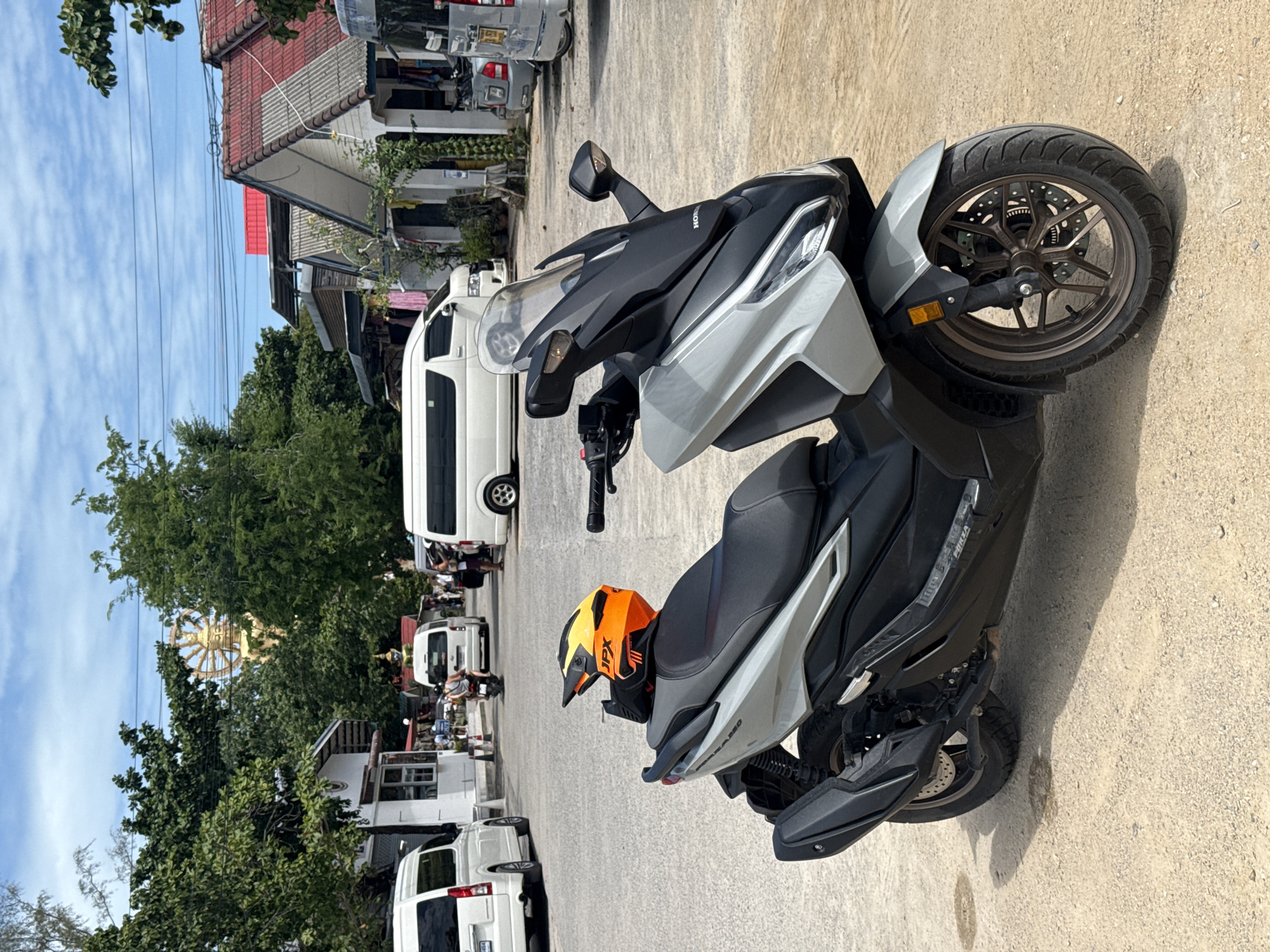 A light gray Honda Forza scooter is parked on a sandy lot in front of buildings under a bright, partly cloudy sky in what appears to be a commercial area in Ko Samui District, Thailand. A bright orange and yellow JPX helmet rests on the scooter's seat. In the background, several white vans are parked, and a golden Buddha statue is visible above the trees. The buildings have red-tiled roofs and some feature white walls. The overall scene suggests a sunny day in a tropical location, likely a tourist area given the presence of vans and the iconic statue.