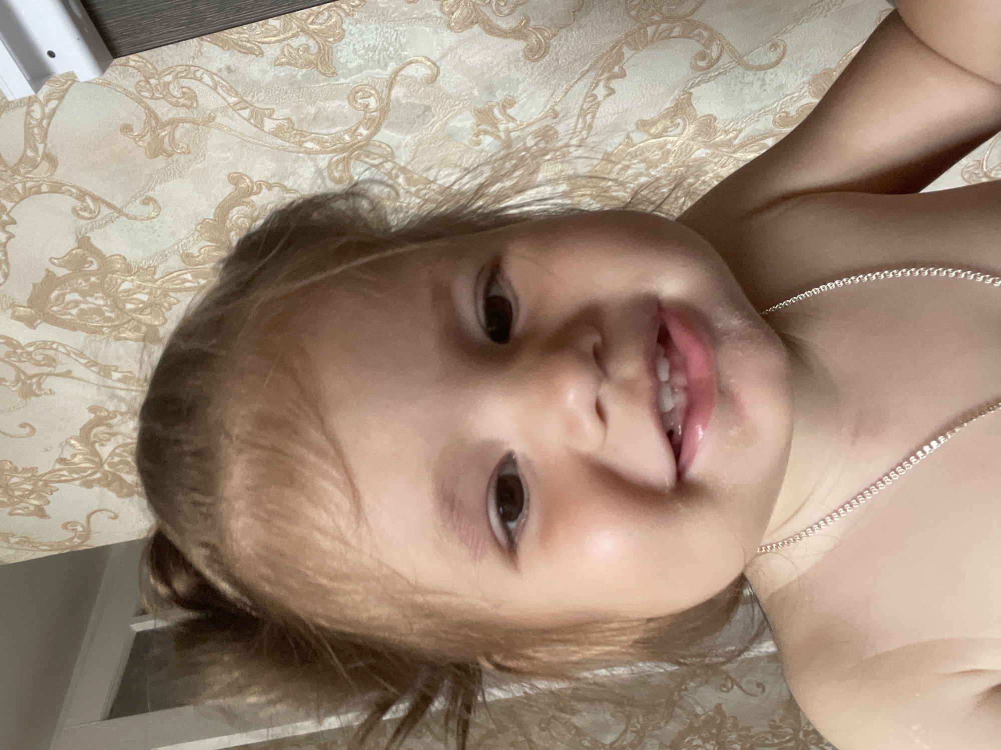 A close-up, waist-up selfie of a young Caucasian child with light brown hair pulled into a ponytail is featured. The child is looking directly at the camera and smiling, showing their teeth. A thin silver chain necklace is visible around the child's neck. The background consists of patterned wallpaper. The image appears to be taken indoors. There are no discernible landmarks or clues to pinpoint a specific location within Izhevsk, Russia.