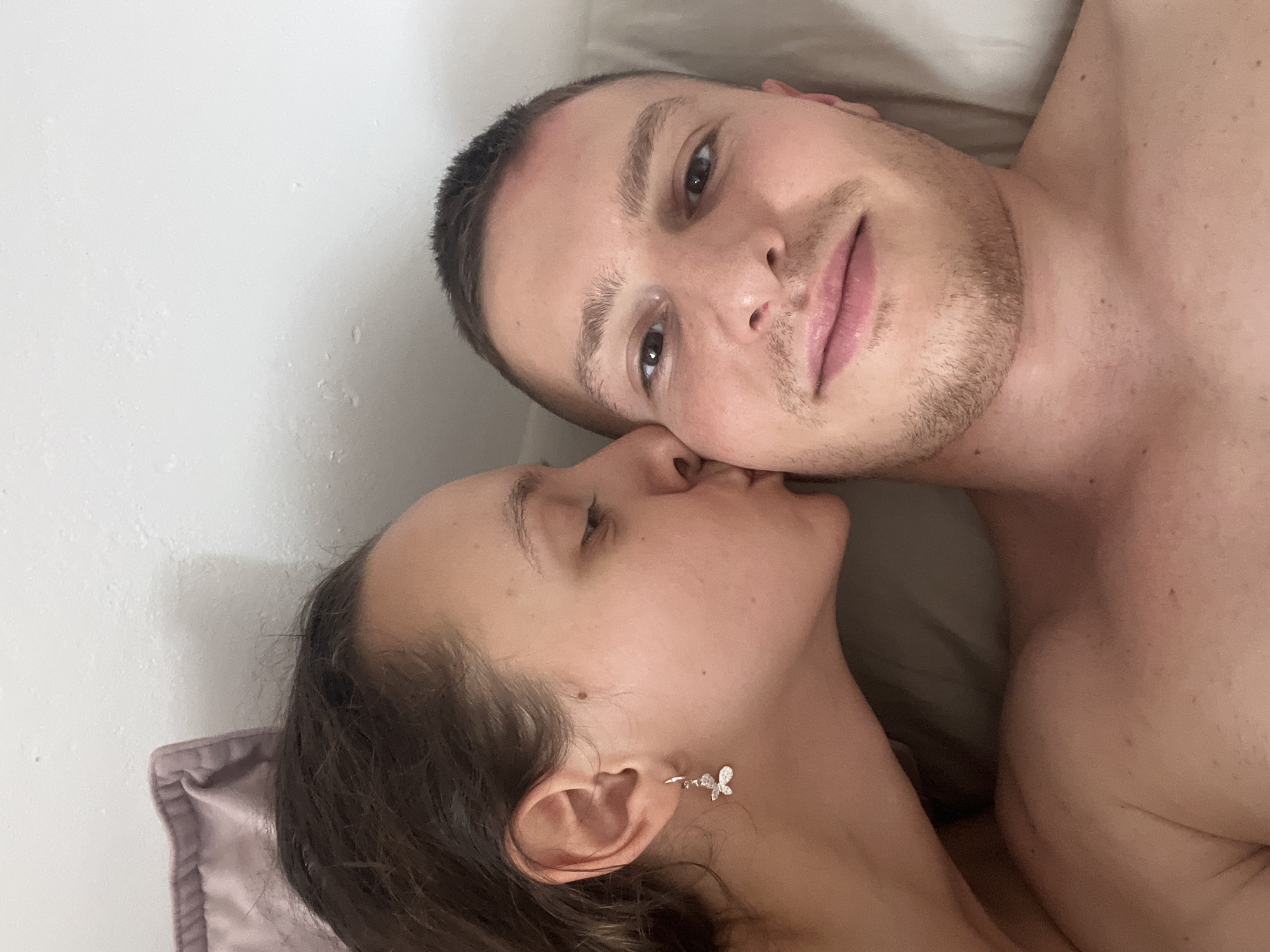 A selfie depicts a man and a woman indoors, lying down. The man is fair-skinned with brown hair and stubble, looking into the camera. The woman, with dark hair and fair skin, is turned toward him, appearing to kiss his cheek. The background suggests a bed or similar soft surface, with a wall visible on one side. There is no indication of this image being in Saarbrücken, Germany.