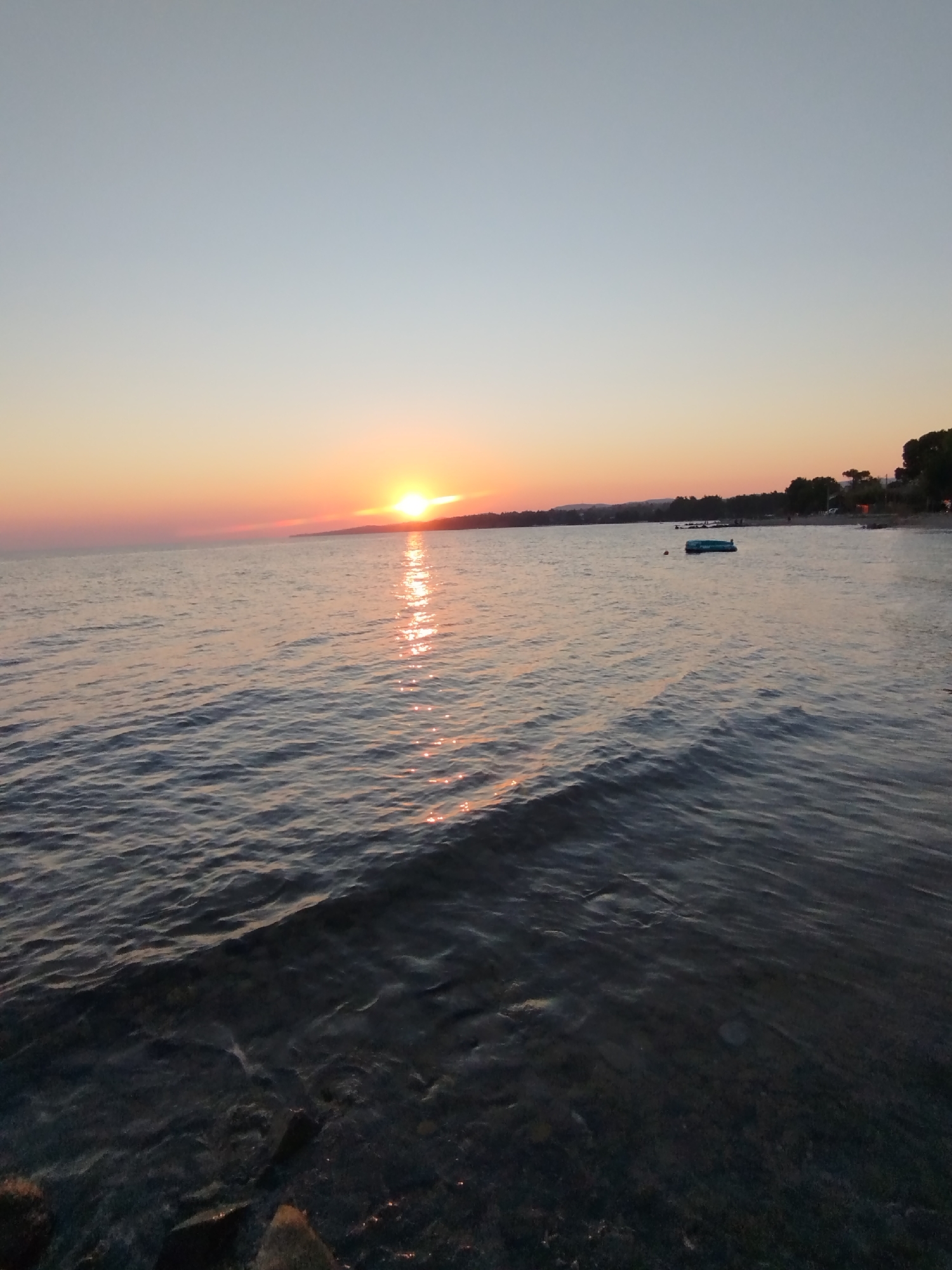 The image depicts a sunset over the sea in Sithonia Municipal Unit, Greece. The sun is low on the horizon, casting a warm glow on the water and the sky, which transitions from pale blue at the top to orange and pink near the horizon. The sea is calm with gentle waves lapping against a rocky shore in the foreground. In the distance, the coastline is visible with a line of trees and some structures, suggesting a populated area. There are a few small objects floating on the water, possibly buoys or small boats, and some indistinct figures can be seen on the shore. The overall atmosphere is tranquil and serene, characteristic of a beach sunset.
