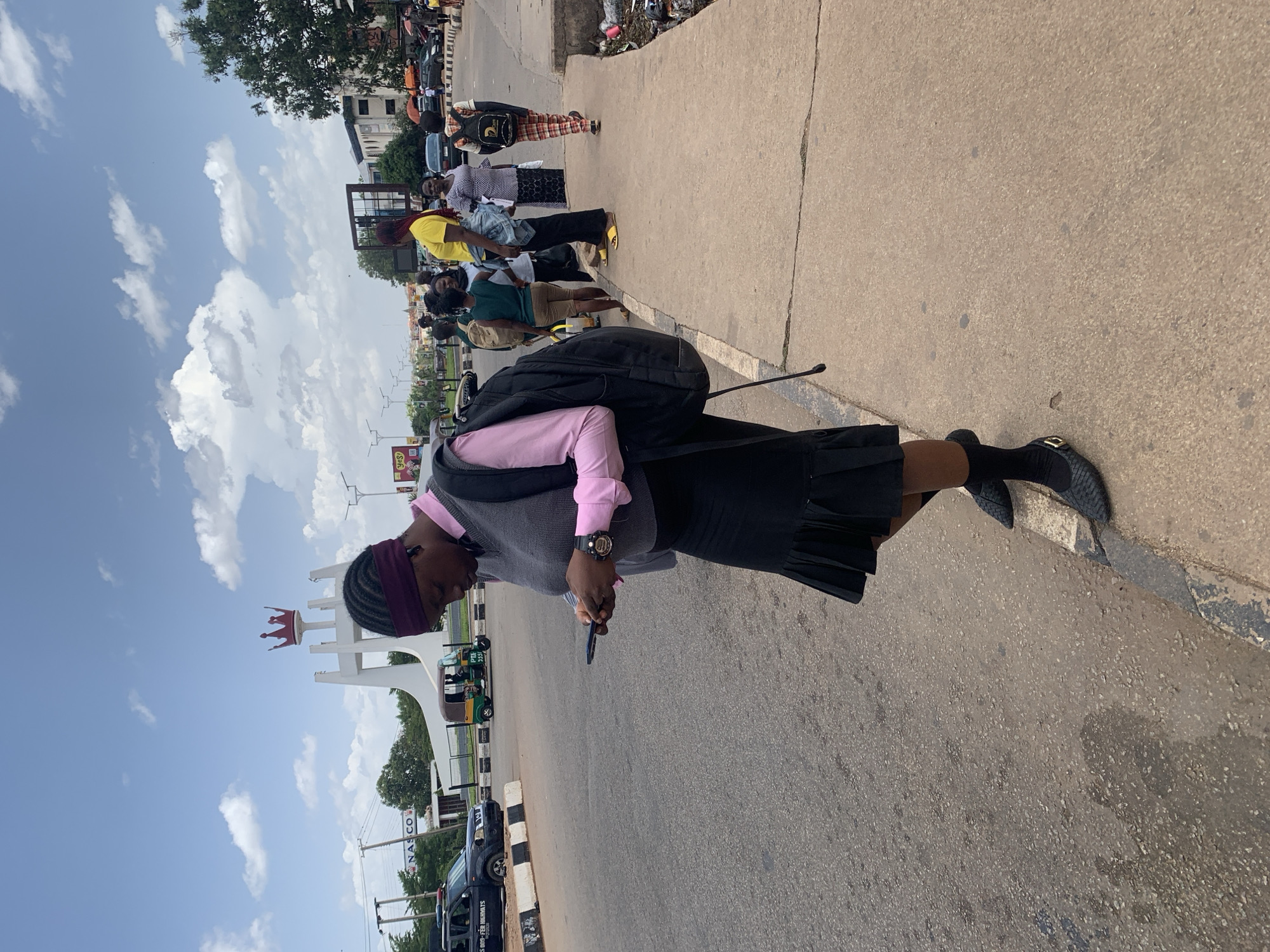 The image captures a daytime street scene in Jos, Nigeria, featuring a woman in the foreground and other people and urban elements in the background under a bright, partly cloudy sky.
The primary subject, seen from a slightly elevated perspective, is a woman standing on a concrete sidewalk. She is dressed in a pink long-sleeved shirt, a grey vest, a black pleated skirt, black socks, and black flat shoes with a decorative buckle. She carries a black backpack and wears a dark red headband over her braided hair. Her gaze is directed downwards at a smartphone held in her hands, and a black wristwatch is visible on her left wrist.
Behind her, several other individuals are visible further down the sidewalk, some appearing to be waiting or walking. They are dressed in casual attire, including one person in a yellow shirt and another in plaid pants.
The scene includes an asphalt road parallel to the sidewalk, separated by a white and black striped curb. Across the road, a prominent white monument topped with a large red crown stands out, a recognizable landmark in the area. Further along the road, a green and yellow tricycle (Keke Napep) and a dark-colored vehicle are visible. Trees and buildings line the street in the distance.
The time of day is clearly daytime, indicated by the bright illumination and the blue sky dotted with scattered white cumulus clouds.
Visible text includes: "PTA 2251" on a yellow and black pole, "NAS CO" on a building in the background, and a partial sign on a black vehicle that reads "THIS SHOP FOR HIGHER UNITS."