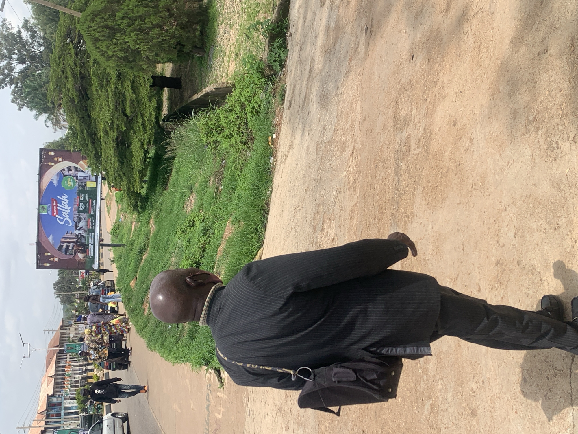 A man in a pinstripe suit and black shoes walks along a dirt path next to a grassy area. He has a black bag slung over his shoulder. In the background, there are trees, buildings, and a billboard. The billboard advertises "Sallah" and displays images of buildings and people. There are also cars and a street vendor with a cart full of goods visible. The scene appears to be a street in Gura Top, Nigeria, possibly during the daytime, given the lighting. The man's posture and the presence of other people suggest a normal day with commercial activity.