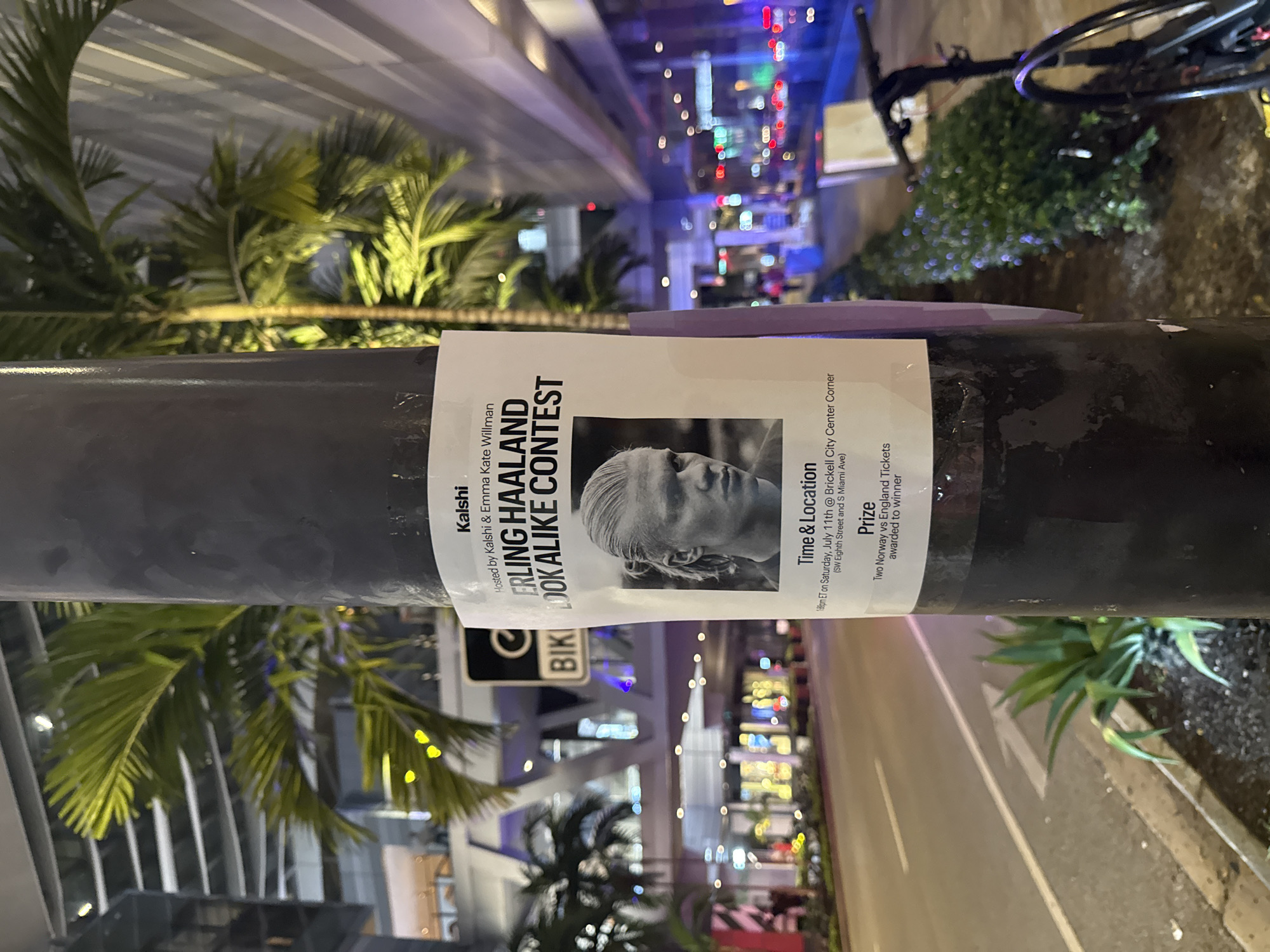 The image shows a poster attached to a lamppost on a city street at night. The poster advertises a "Erling Haaland Look Alike Contest" hosted by Kalshi and Emma Kate Willman. It includes a black and white photograph of Erling Haaland, a soccer player. The poster also lists the time and location of the event: 7:00 PM ET on Saturday, July 11th, at the Brickell City Center Corner (SW Eighth Street and S Miami Ave). The prize is stated as "Two Norway vs England Tickets awarded to winner."
In the background, there are palm trees, modern buildings with illuminated windows, streetlights, and traffic lights, indicating an urban environment. The street appears to be wet, possibly from rain or recent watering. A bicycle is partially visible on the right side of the image, parked near some bushes. The overall scene suggests a lively urban setting, likely in Miami, given the mention of Brickell and Miami Ave.