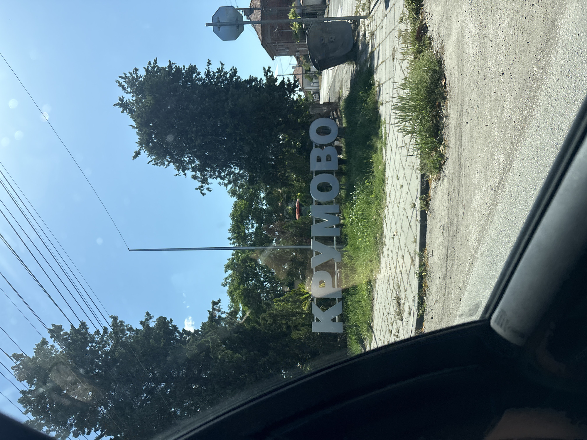 The image shows the entrance sign to the city of Krumovo, Bulgaria. The sign is made of large, white letters spelling out "КРУМОВО" (Krumovo in Cyrillic). The sign is located on a paved area near a road and sidewalk. There are trees and greenery behind and around the sign. A utility pole with a traffic signal or sign is also visible near the sign. The image was taken from inside a vehicle, with part of the car's interior visible in the foreground. The sky is blue and the lighting suggests it is daytime. Buildings are visible further back in the shot, providing context to the location within the city.
