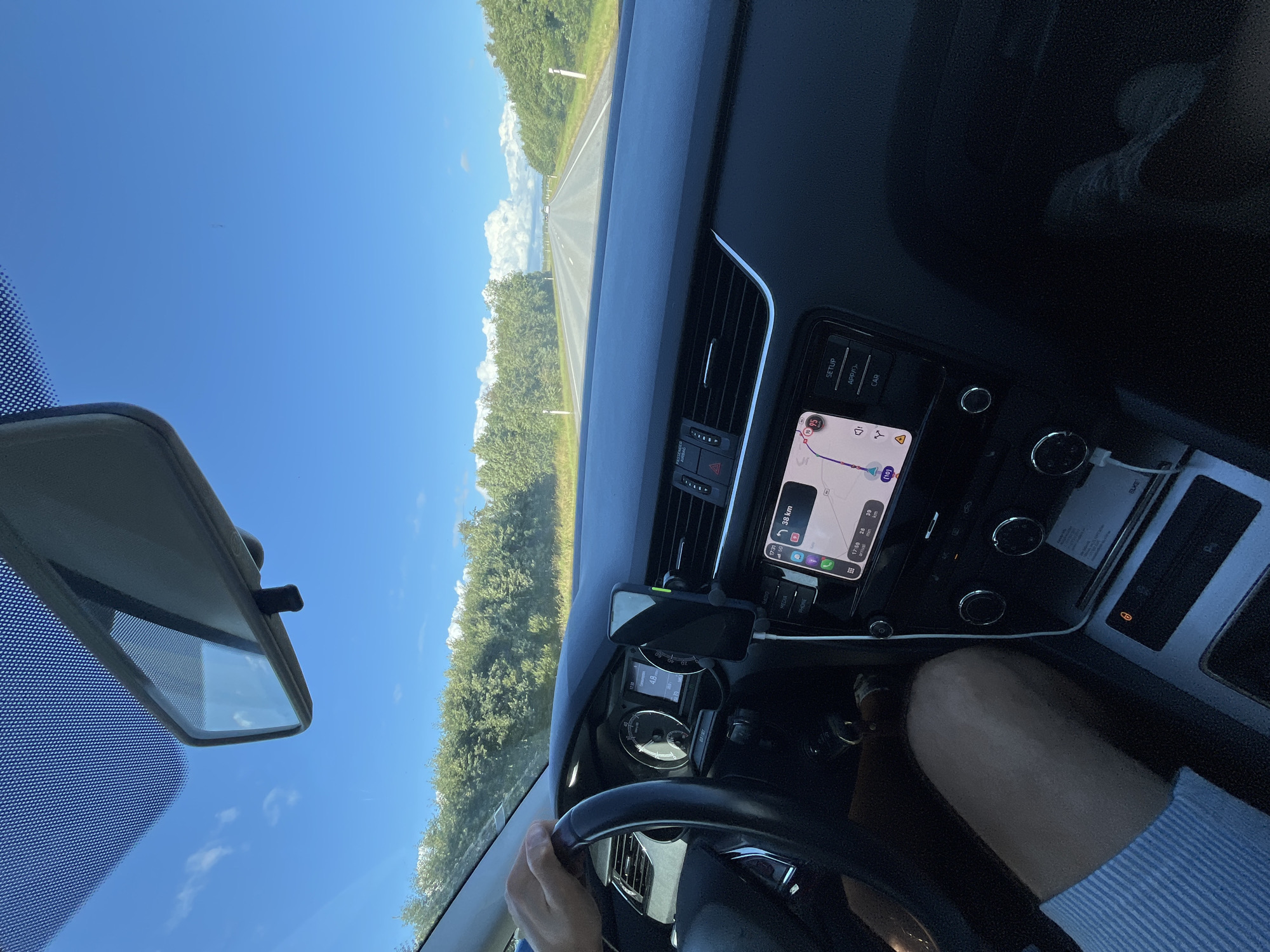 The image depicts the interior of a car in motion on a road. The driver's hand is visible on the steering wheel, and their legs are also in view. A smartphone is mounted on the dashboard, displaying navigation information with a route highlighted. The car's dashboard features a digital display showing speed and other vehicle information. Outside the car, a tree-lined road stretches into the distance under a clear blue sky with scattered clouds. The location is identified as Lihula, Estonia.
