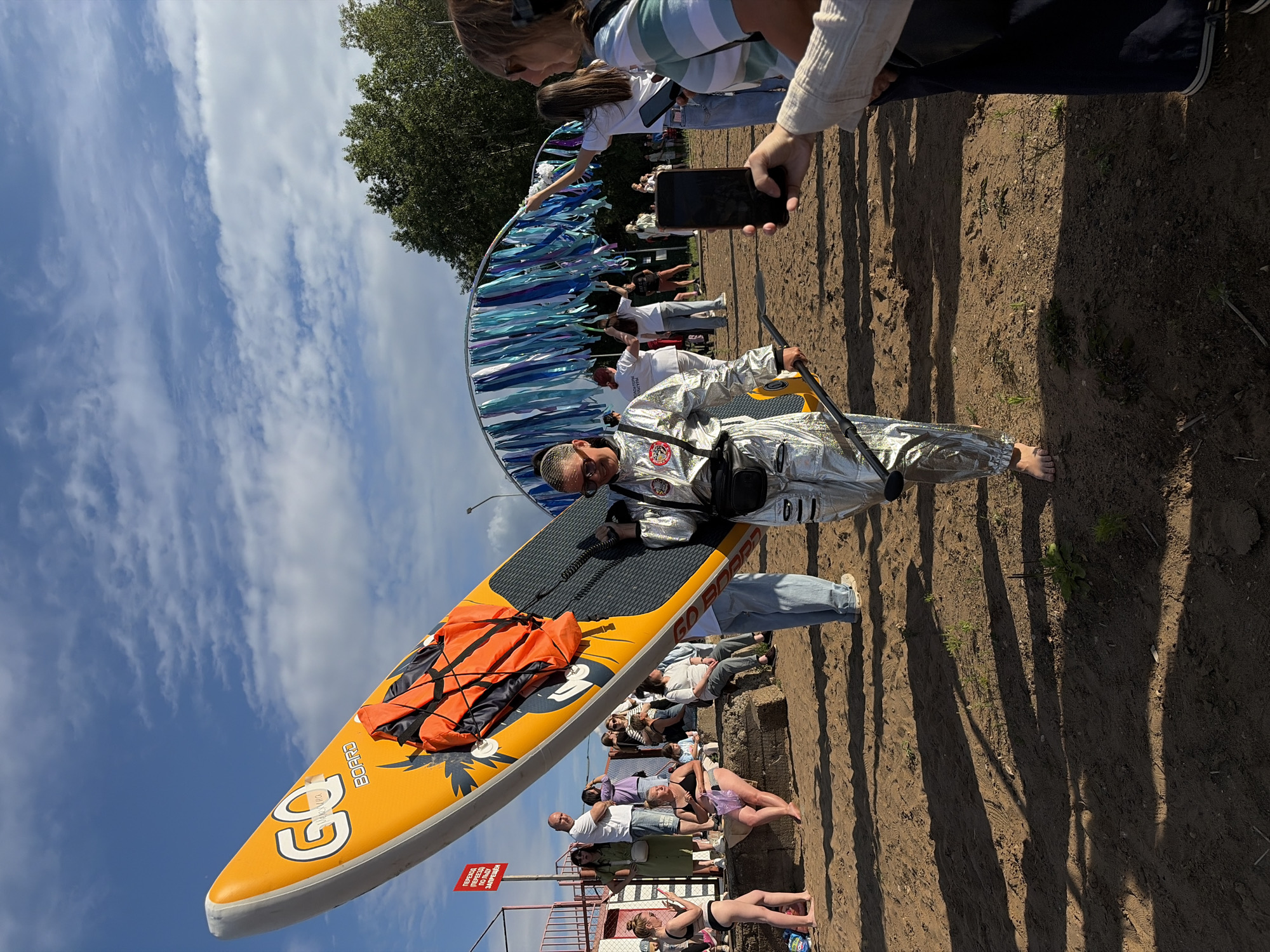 A person in a silver, full-body, astronaut-like suit walks on a sandy beach with a bright orange and yellow paddleboard held vertically. The paddleboard has "GO BOARD" printed on it and is equipped with an orange life vest secured to it with straps. The person is barefoot and carries a black paddle in their right hand, with a black fanny pack worn across their chest. In the background, a group of people are gathered on a stepped embankment, some sitting and others standing. A decorative archway draped with blue and white ribbons is visible behind the crowd. The environment appears to be a public beach with trees and a blue sky with scattered clouds. The image is taken in Chaikovsky, Russia, suggested by the presence of the Kama River and its surrounding environment, typical of recreational areas in the city.