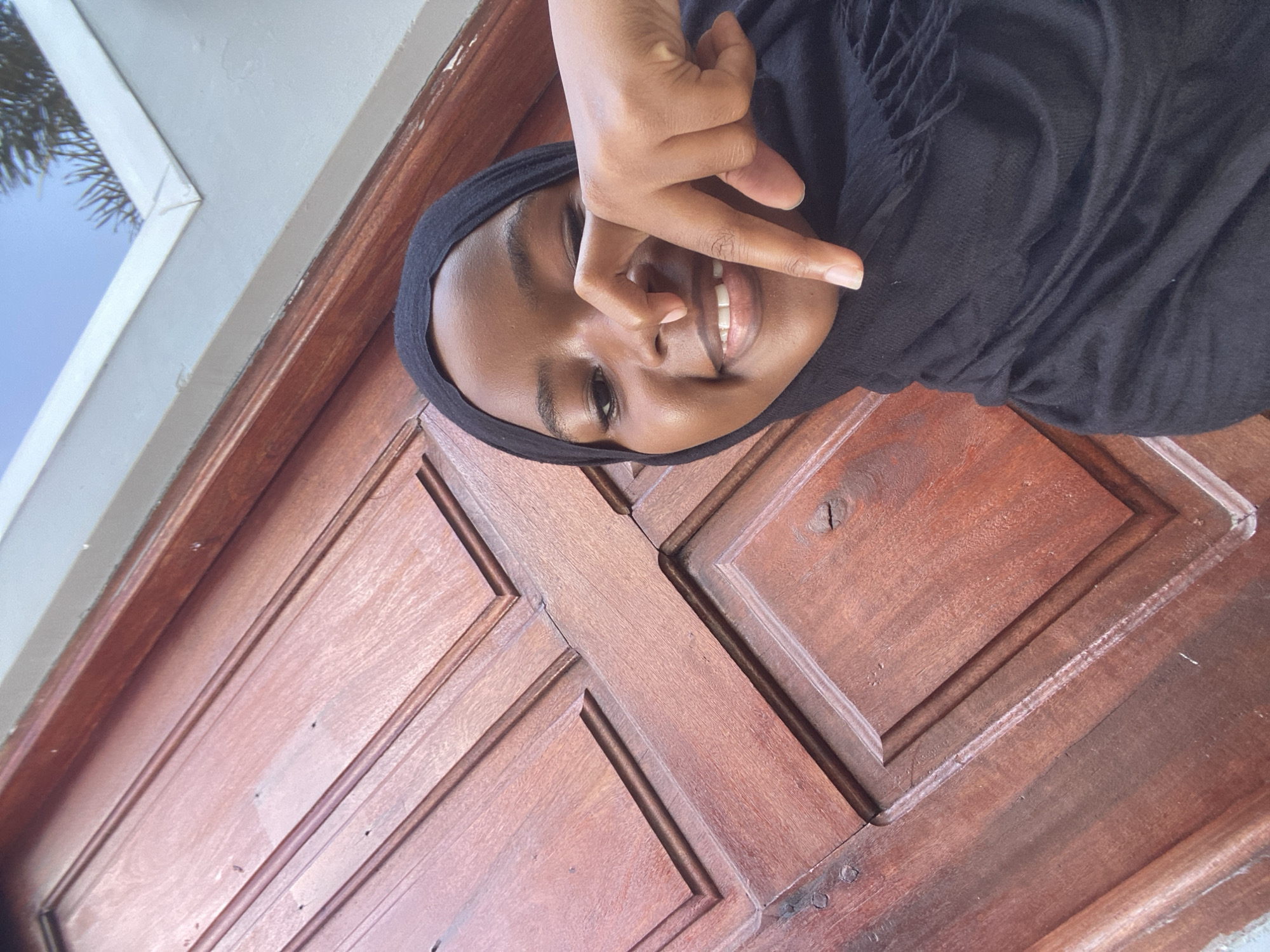 An image taken outdoors or semi-outdoors depicts one young adult female, dark-skinned, smiling, and looking directly at the viewer. She is wearing a black headscarf (hijab) and a black garment. Her right hand is raised, with the index finger near her nose. The primary background consists of a reddish-brown wooden door or paneling with recessed panels and carved decorative mouldings. To the right of the subject, a light-colored wall or frame is visible, beyond which a clear blue sky and green foliage are discernible. The exact location within Dar es-Salaam, Tanzania, cannot be determined from the visible elements.