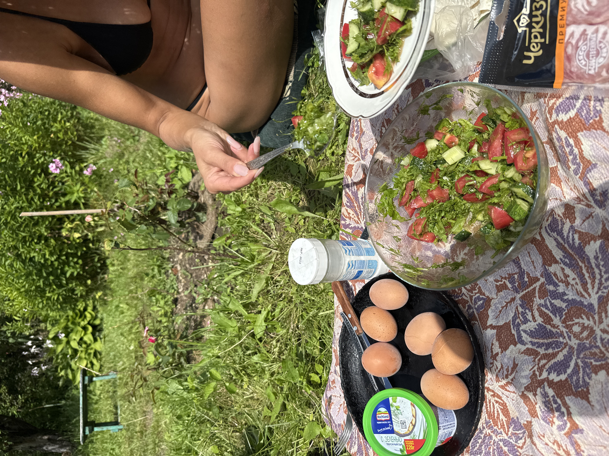 A high-angle medium shot captures a picnic scene on a sunny day in a grassy area, possibly a garden or park. In the foreground, a colorful floral-patterned blanket is spread on the ground. Several raw eggs are arranged on a black circular dish. To the left of the eggs, a green tub of what appears to be cream cheese or a spread is visible, with some text in Cyrillic. A salt shaker with a white lid stands next to the eggs. A clear glass bowl filled with a chopped salad of tomatoes and cucumbers, mixed with herbs, is positioned in the center of the blanket. A fork rests on the blanket near the bowl. In the middle ground, a white plate holds a portion of the same salad. To the right of the plate, a package of sliced meat with Cyrillic labeling, possibly from "Cherkizovo," is partially visible. A person, seen from the waist down, is sitting on the blanket, wearing a black bikini top. Their hand is holding a spoon, scooping a portion of salad from the glass bowl. The background shows lush green grass and foliage, with a hint of pink flowers. In the further background, a rustic green bench is visible amongst the greenery, and a dense thicket of green bushes and trees forms the backdrop. The sky is not visible in the frame. The lighting suggests it is daytime, with bright sunlight casting shadows.