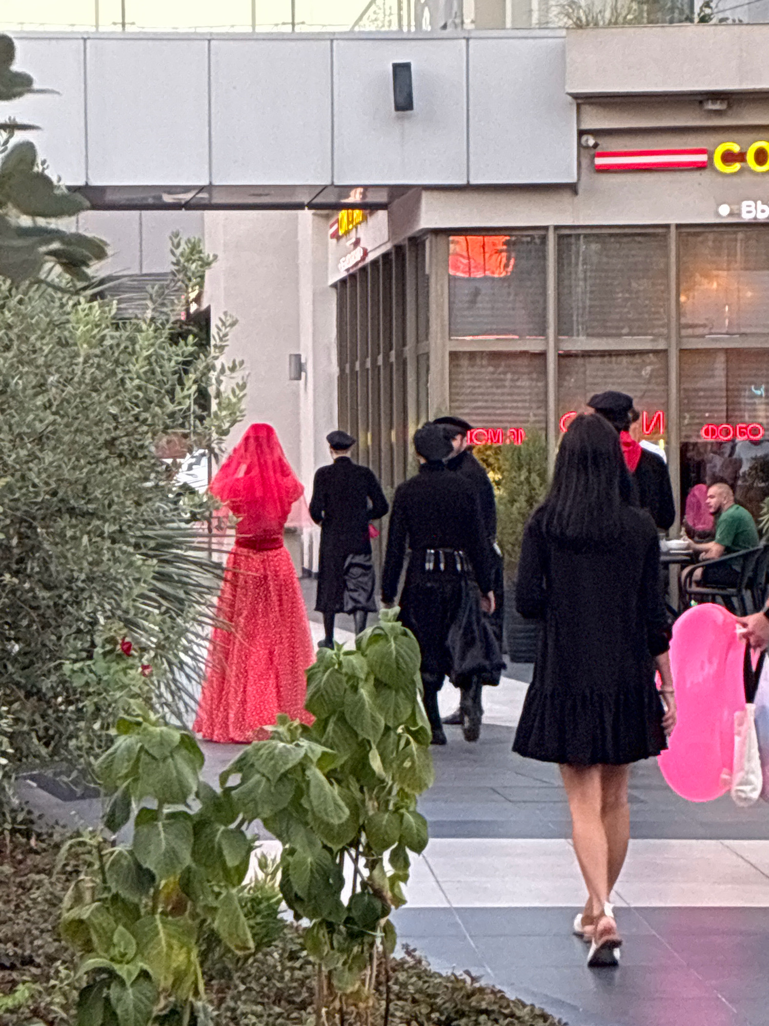 In Sochi, Russia, on an outdoor pedestrian walkway, a group of people dressed in traditional attire is walking away from the camera. On the left, a person in a red, textured dress and a red veil is prominent. To their right, several individuals are wearing black, floor-length coats, black hats, and boots, suggesting a cultural event or performance. In the background, a building with large windows displays neon signage, partially obscured by reflections. One sign appears to read "CO" in yellow letters. A man in a green shirt is seated at a table outside the building, facing away from the camera. A large, bright pink object, possibly a flotation device, is carried by someone in the foreground on the right. Lush green plants are visible in the foreground, framing the scene.