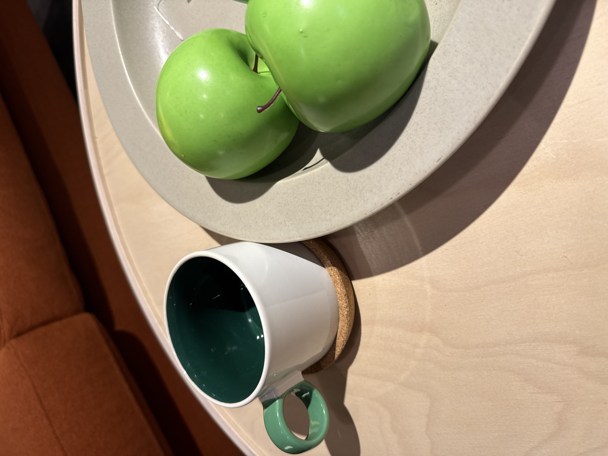 A high-angle close-up shot shows a light-colored wooden surface with a gray plate holding two green apples. A white mug with a green interior and handle sits on a cork coaster beside the plate. Part of an orange sofa is visible in the lower left corner. The scene is likely indoors, focusing on a still life arrangement of the mentioned objects. There is no evidence to suggest this image was taken in Sofia, Bulgaria.
