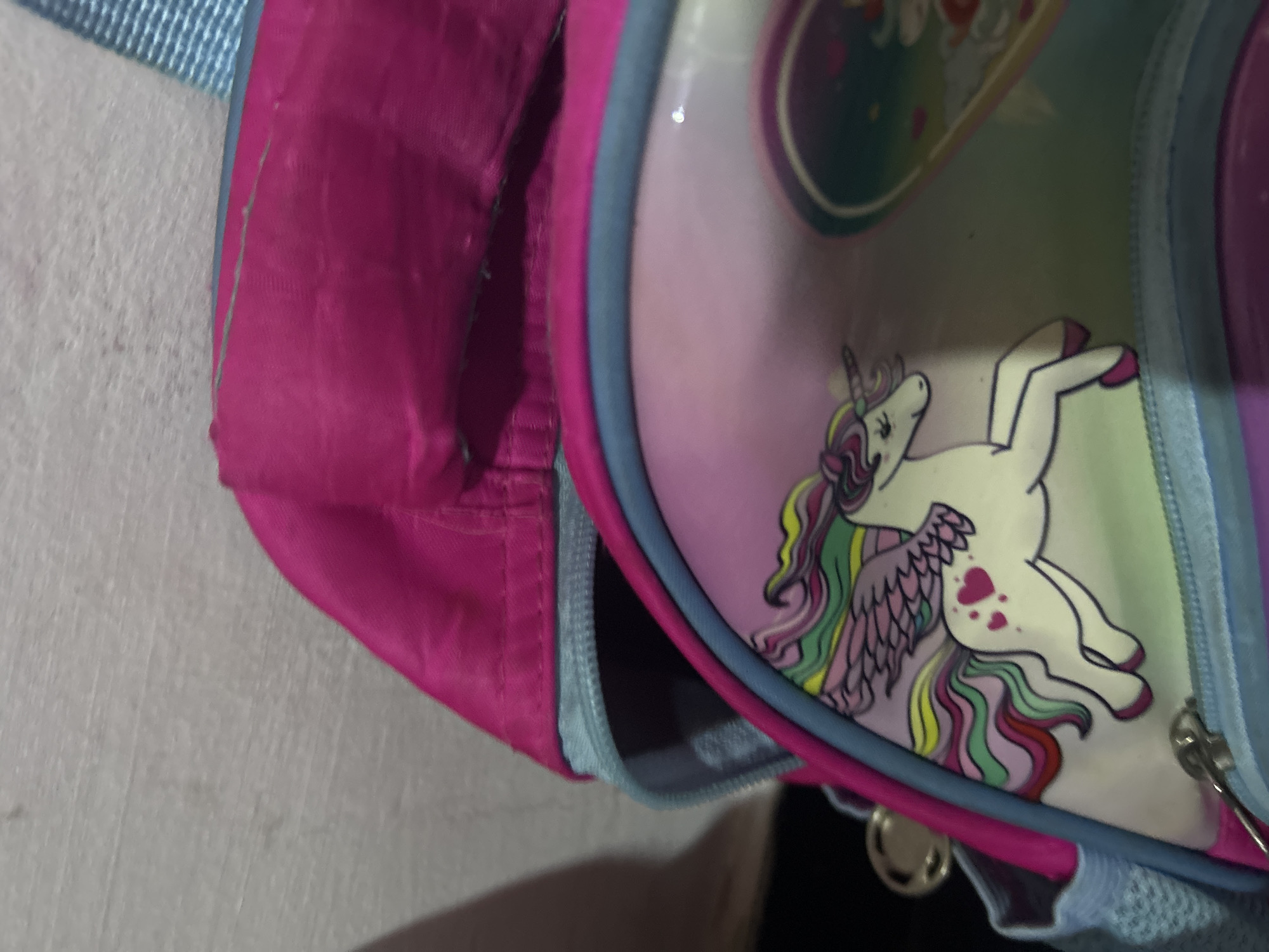 A close-up, high-angle shot shows part of a pink and light blue child's backpack featuring a unicorn design. The backpack is comprised of a pink fabric pocket section on the left and a rounded, transparent plastic section on the right, showcasing a white unicorn with a multicolored mane and hearts on its body. The unicorn is lying down within a pastel-colored design. A light blue zipper runs along the perimeter of the plastic section, with a zipper pull visible. The background is blurry and shows a light-colored surface on the left and a dark-colored area below the backpack. There is no indication of the location being San Juan de Los Morros, Venezuela.