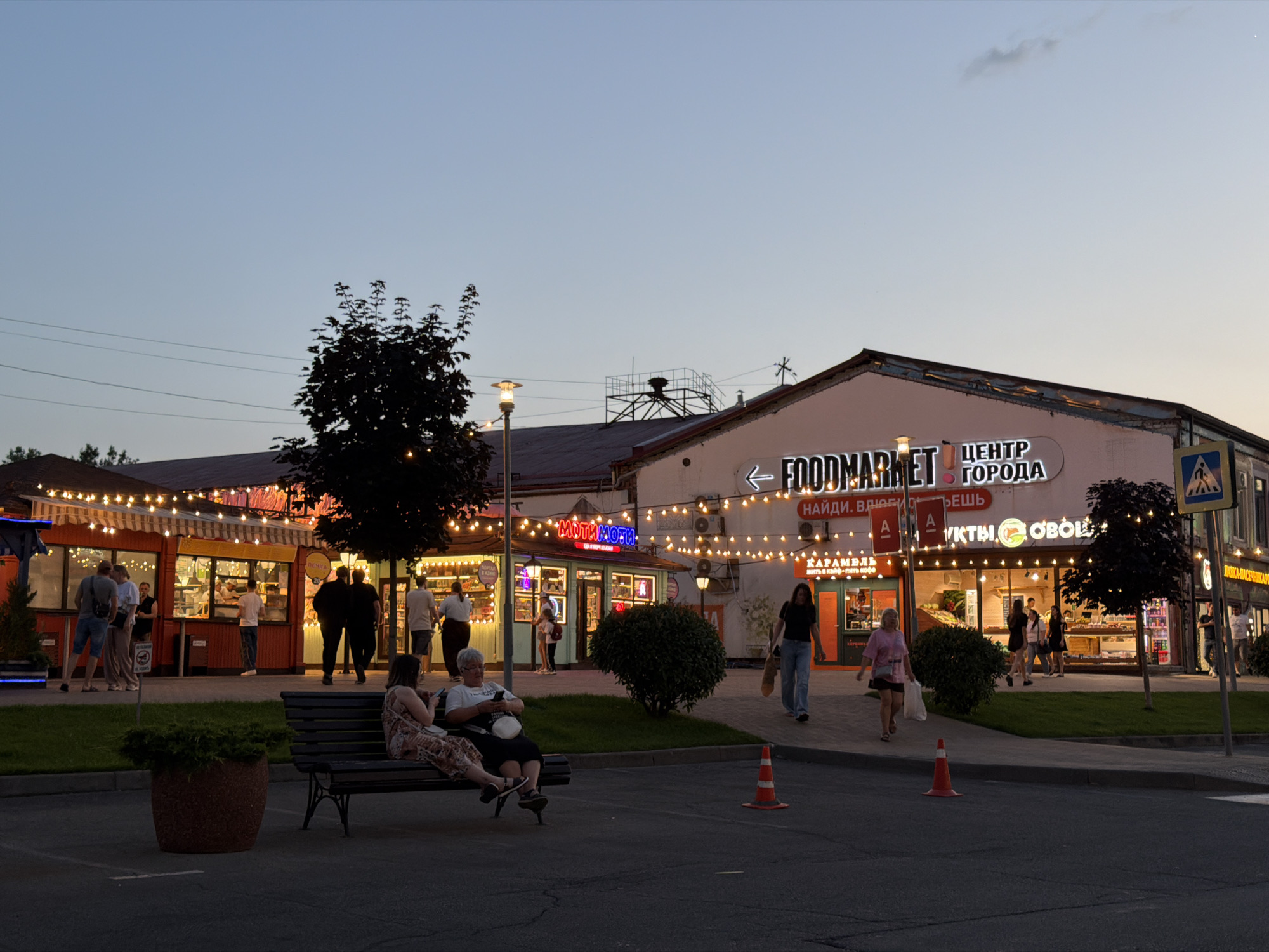 The image captures a bustling outdoor market area at dusk, illuminated by strings of warm-toned lights. Located in Krasnodar, Russia, specifically at the Foodmarket city center, the scene showcases various food stalls and shops. In the foreground, a dark-colored bench holds two women, one seated and the other leaning back, both appearing relaxed and engaged in conversation.
Further back, a group of people are seen standing and walking around the market stalls. Some are browsing the merchandise, while others are conversing or moving through the area. The stalls themselves are brightly lit, displaying a variety of goods, including what appears to be produce and baked items. The architecture of the market features a long, low building with a pinkish facade, adorned with neon signs and the prominent "FOODMARKET" lettering.
The environment suggests a pleasant evening atmosphere, with a clear sky overhead and well-maintained pathways and landscaping. Several trees, including a large dark-leaved tree on the right, contribute to the outdoor setting. Orange traffic cones are visible on the pavement, indicating some form of temporary closure or demarcation. A street sign with a blue and yellow pedestrian crossing symbol is also present, suggesting proximity to a road. The overall impression is one of a vibrant and active community gathering space.