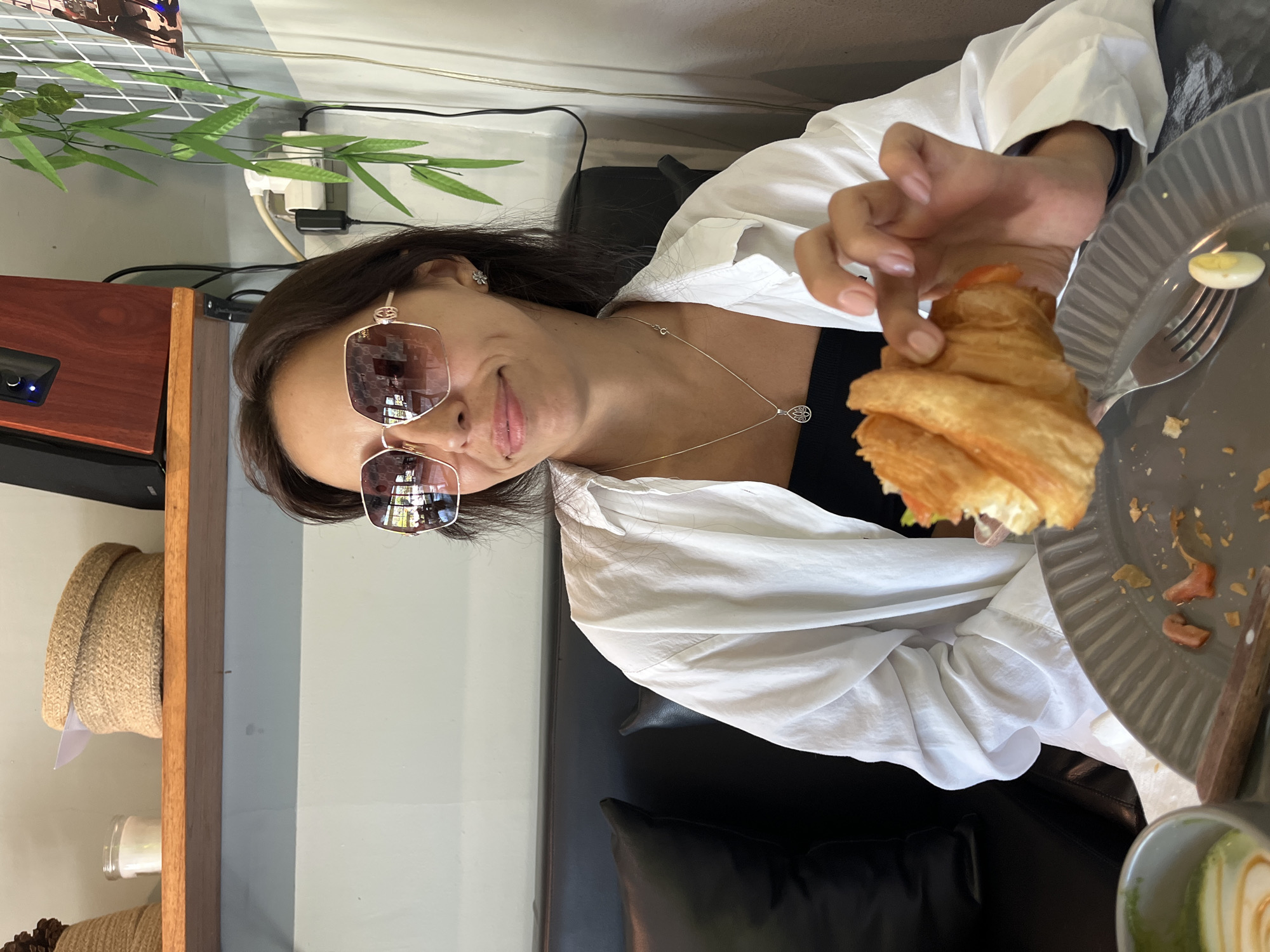 The image captures a woman indoors, likely in a café or restaurant setting. She is wearing sunglasses and a white shirt, holding a croissant in her hand near a plate with a fork and what appears to be a halved egg. A portion of a dark-colored sofa or chair is visible behind her. In the background, there is a decorative shelf with various items and a potted plant. The photograph was taken in Kuta Selatan, Indonesia.