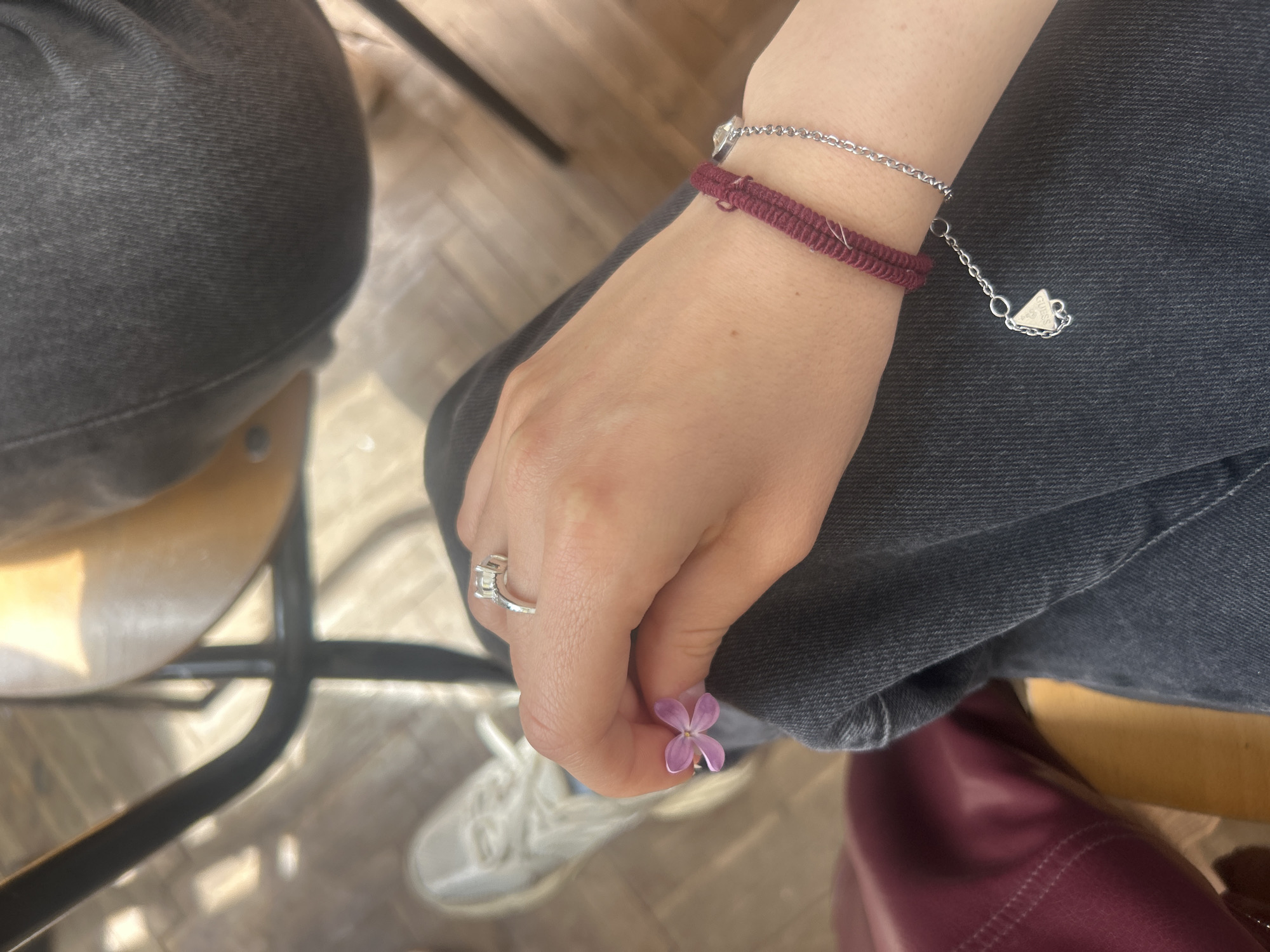A top-down, medium shot depicts a person's hands, a small flower, and jewelry in an indoor setting.
The person is seated on a chair with black metal legs. They wear dark gray jeans and a pair of white sneakers. Their right hand is visible, holding a tiny, purple flower with four petals between their thumb and forefinger. On the ring finger of their right hand, they wear a silver-colored ring with a prominent stone setting. Two bracelets adorn their right wrist: a braided, burgundy cord bracelet and a delicate silver chain bracelet with a "Guess" pendant. A portion of a burgundy-colored leather bag is visible to the right. The background features wooden flooring. The image appears to be taken during the daytime under natural lighting.