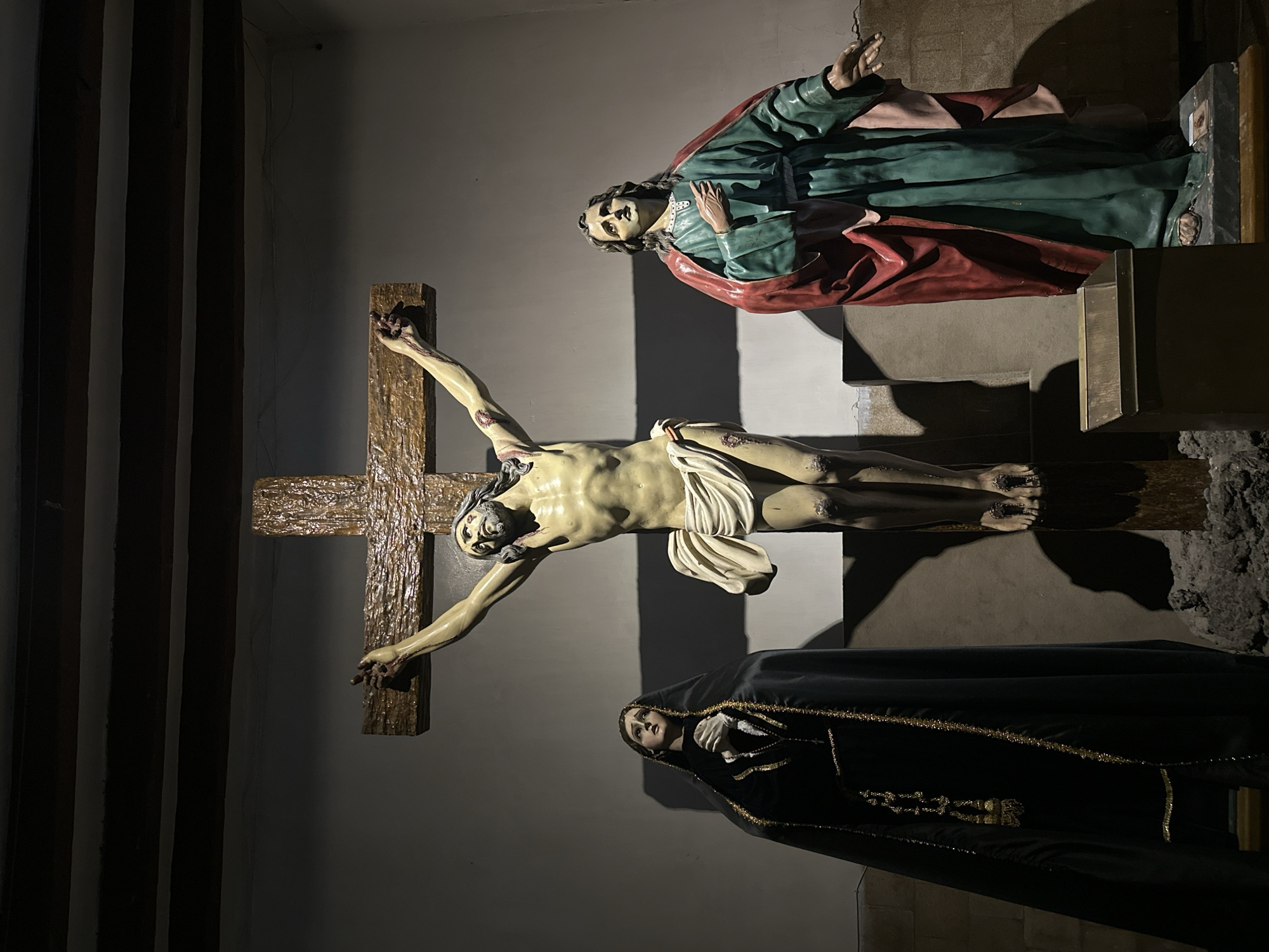 The image depicts a religious scene, likely within a church or cathedral in Mexico City, Mexico, featuring statues arranged to represent the crucifixion of Jesus Christ. The central figure is a statue of Jesus on the cross, with arms outstretched and a white cloth draped around his waist. Above him stands a statue of a man with dark hair, wearing a green and red robe. Below the crucified Jesus, a statue of a woman, possibly Mary, is present, draped in a black robe with gold trim, her face expressing sorrow or mourning. The background is a dimly lit wall, and the overall composition suggests a somber and reverent atmosphere.