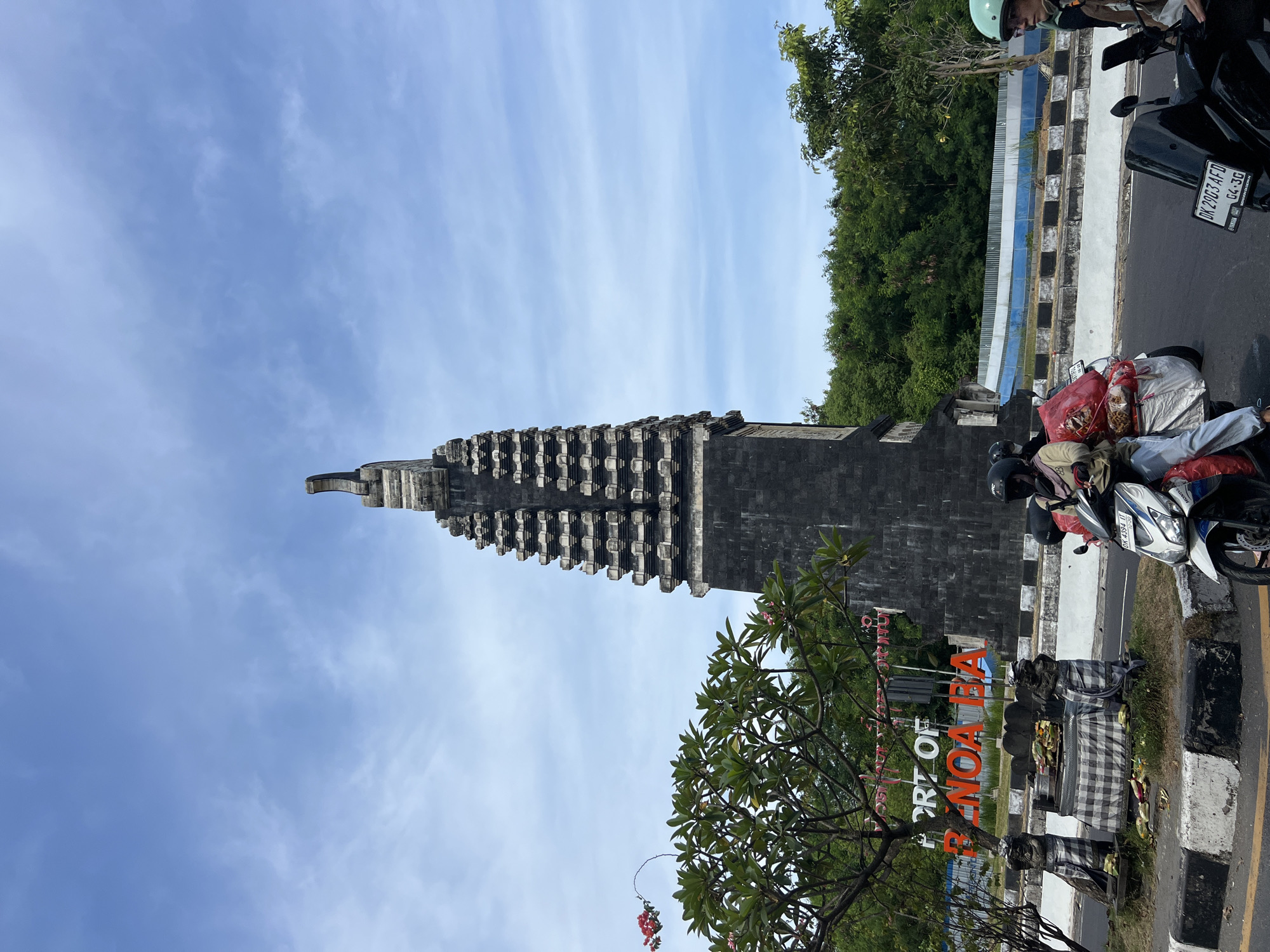 A stone monument with a tiered design stands against a bright blue sky streaked with wispy clouds. Adjacent to the monument is a sign that reads "PORT OF BANOA BA". A tree with green leaves and red blossoms partially obscures the view of the sign. Parked on the side of the road are two motorbikes, one black and one white, loaded with bags and objects, one showing the plate number "DK 2903 AFD". A person wearing a light green helmet is positioned on one of the motorbikes. In the background, dense green trees line a fence. The setting appears to be in the Denpasar, Indonesia, region.