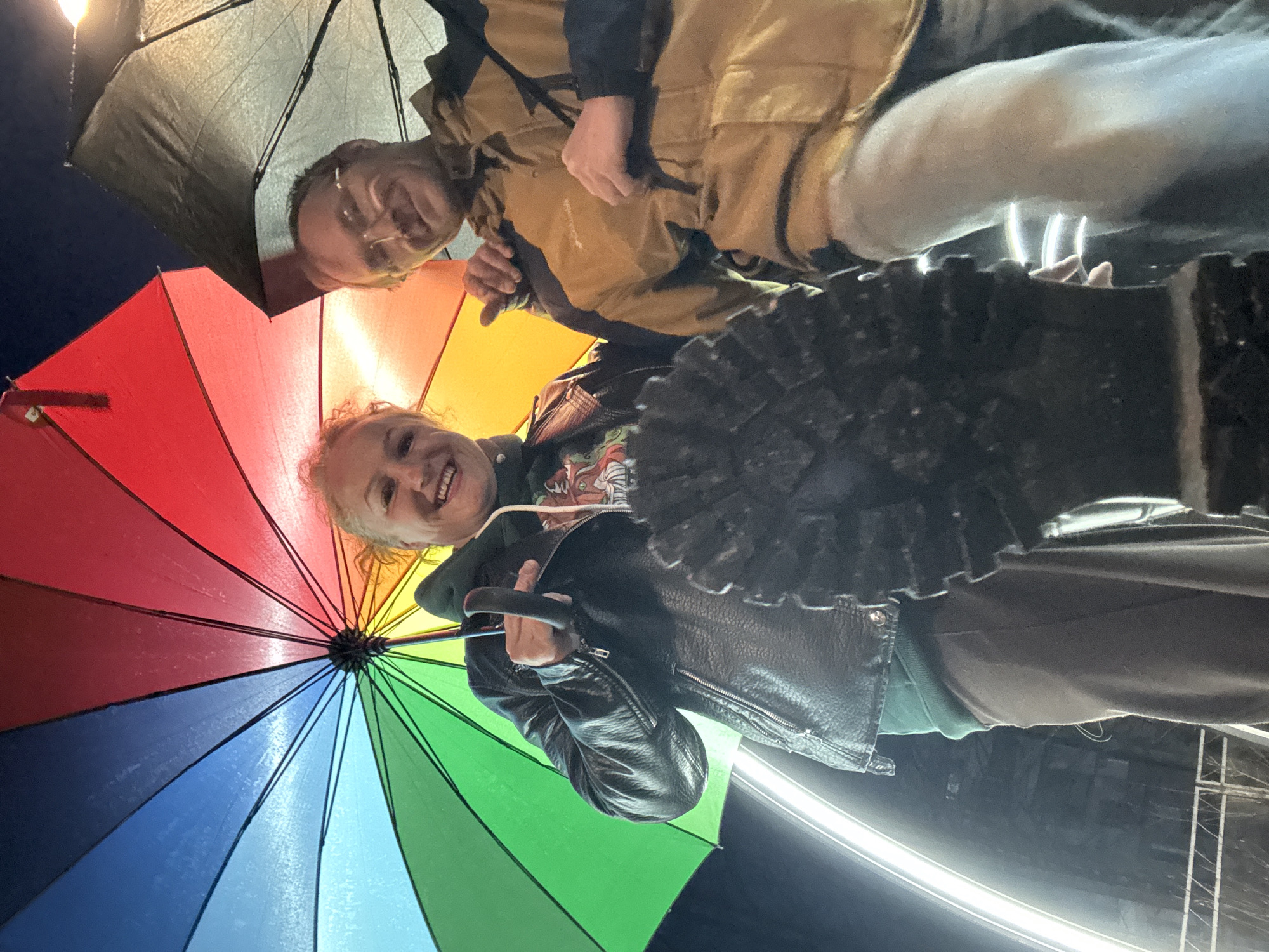 Two people stand under umbrellas in the city of Chaikovsky, Russia. One umbrella is rainbow-colored, while the other is grey. The first person wears a black leather jacket. The second person wears a tan jacket and glasses. One person points upward towards the camera. The image is taken from a low angle, showing the bottom of a boot. Bright white lights are in the background.