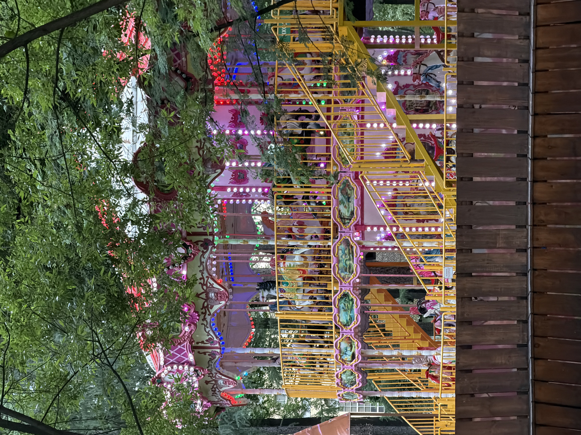 The image shows a carousel at what appears to be a fairground or amusement park, possibly during an evening event, given the illuminated lights. The carousel is decorated with colorful lights, primarily pink, purple, and red, which are strung along its structure and also form patterns on the ride itself. The carousel has multiple levels and features a yellow metal staircase that provides access to the upper decks. The horses and other figures typically found on a carousel are visible, some decorated with detailed paintings. Surrounding the carousel, trees with lush green foliage partially obscure the view, suggesting an outdoor setting with natural elements. To the right of the carousel, there is a wooden structure, possibly a fence or a booth, made of horizontal planks. The lighting suggests it might be dusk or night, enhancing the vibrancy of the carousel lights. The overall scene conveys a festive and lively atmosphere.