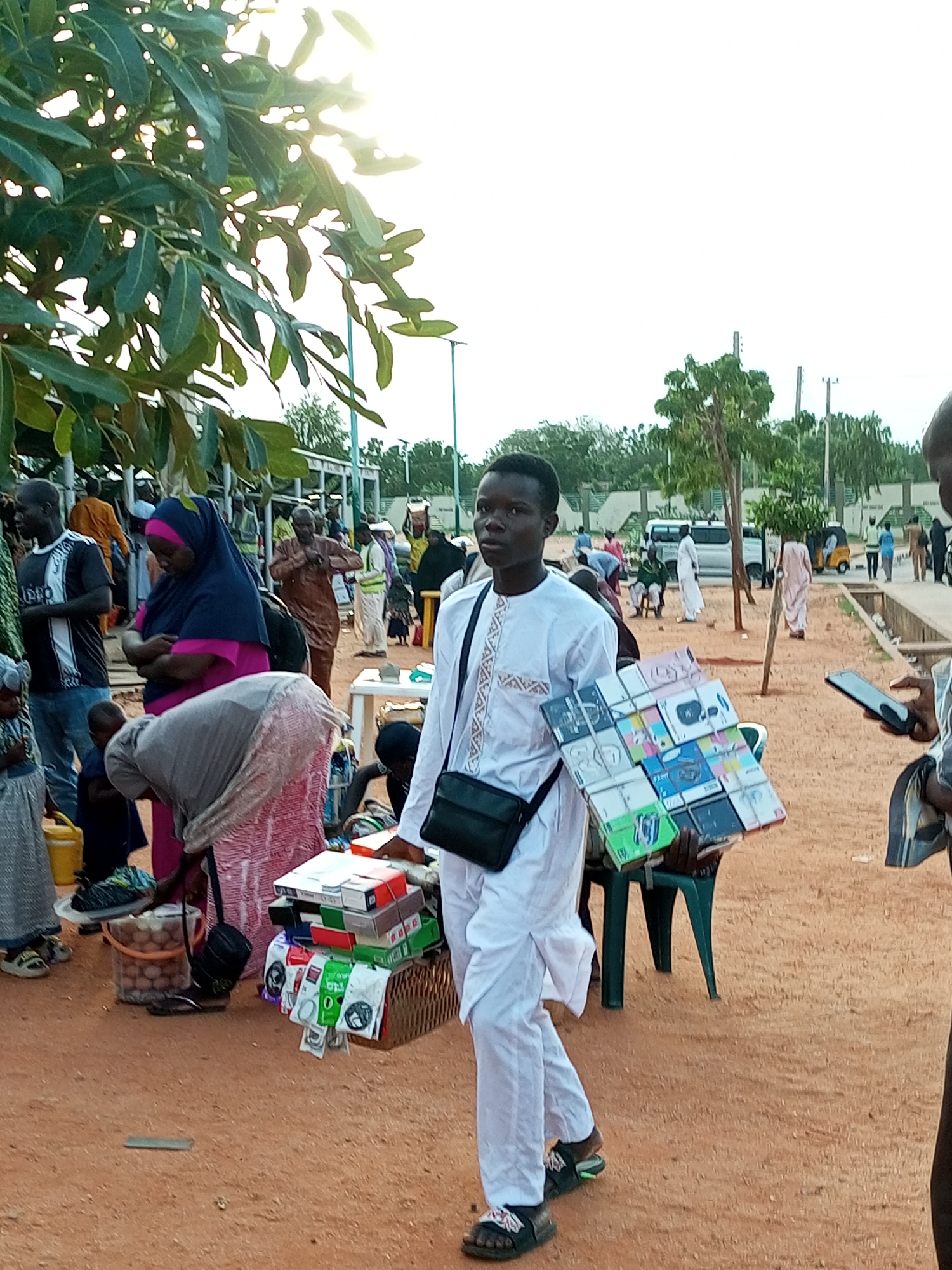 A medium shot captures a busy outdoor market scene in Gombe, Nigeria, with a bright, overcast sky and scattered trees providing dappled shade. In the foreground, a young man dressed in a white traditional outfit walks towards the viewer, holding a flat, square display of various items, possibly phone accessories. He wears a black fanny pack across his chest and black sandals. Behind him, a diverse crowd of people is milling about.
To the left, a woman in a blue and pink hijab stands with her arms crossed, partially obscured by another person. Further left, a man in a black and white t-shirt is visible. In the mid-ground, several individuals are browsing goods displayed on tables and baskets. Among these items are stacks of small boxes, some featuring colorful packaging, and what appear to be packets of accessories. A yellow jug sits near the ground to the left.
In the background, more people are visible, including a man in a brown traditional robe. Beyond them, a white van and a three-wheeled vehicle (likely a "Keke Napep") suggest transportation is present. The ground is composed of dry, sandy earth, and a low wall or barrier is visible in the far background. The overall atmosphere suggests a lively commercial environment.