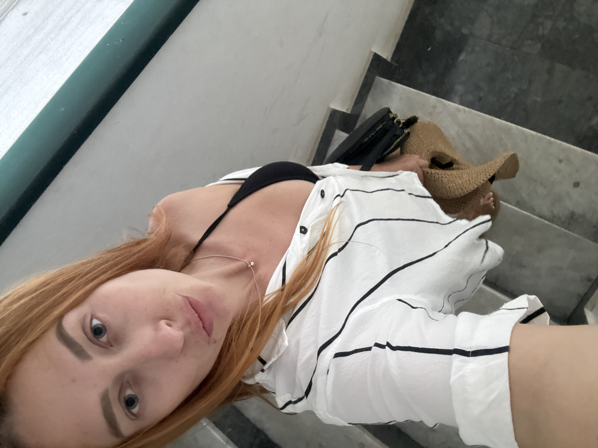 A woman with long auburn hair and light blue eyes, wearing a black bikini top and an unbuttoned white shirt with black vertical stripes, stands on a marble staircase. She is holding a straw hat in her right hand, and a black purse rests on the step beside her. The background shows a green railing along the wall. The scene is likely on the steps of a building in Budva, Montenegro, given the marble construction and the presence of a railing, common architectural features in coastal Mediterranean cities.