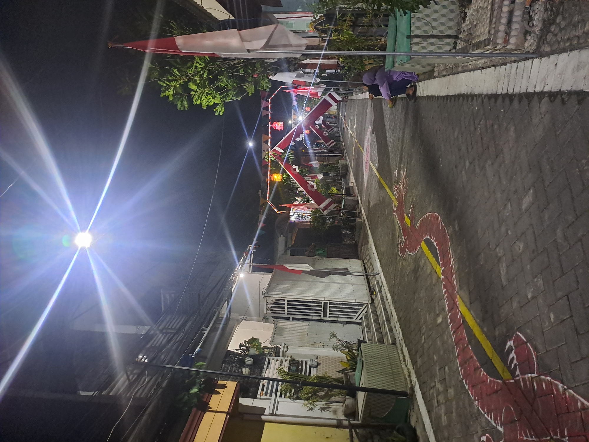 The image depicts a street scene at night in Kebonagung, Indonesia, illuminated by a bright overhead light and some decorative lanterns. The street is paved with dark bricks and features a yellow line with a red, serpentine design painted along its edge. Several Indonesian flags, red and white, are prominently displayed, some attached to poles and others forming part of an arched decoration spanning the street. Small triangular flags in red and white are strung across the scene as well.
On the right side of the street, two individuals, one wearing a purple hijab, are seated on a low wall. The environment includes houses with barred gates and windows, potted plants, and what appears to be some form of decorative paving or structure on the right edge. The overall impression is of a street decorated for a celebration or national event. The lighting creates strong lens flares and highlights, indicating a nighttime setting with artificial light sources.