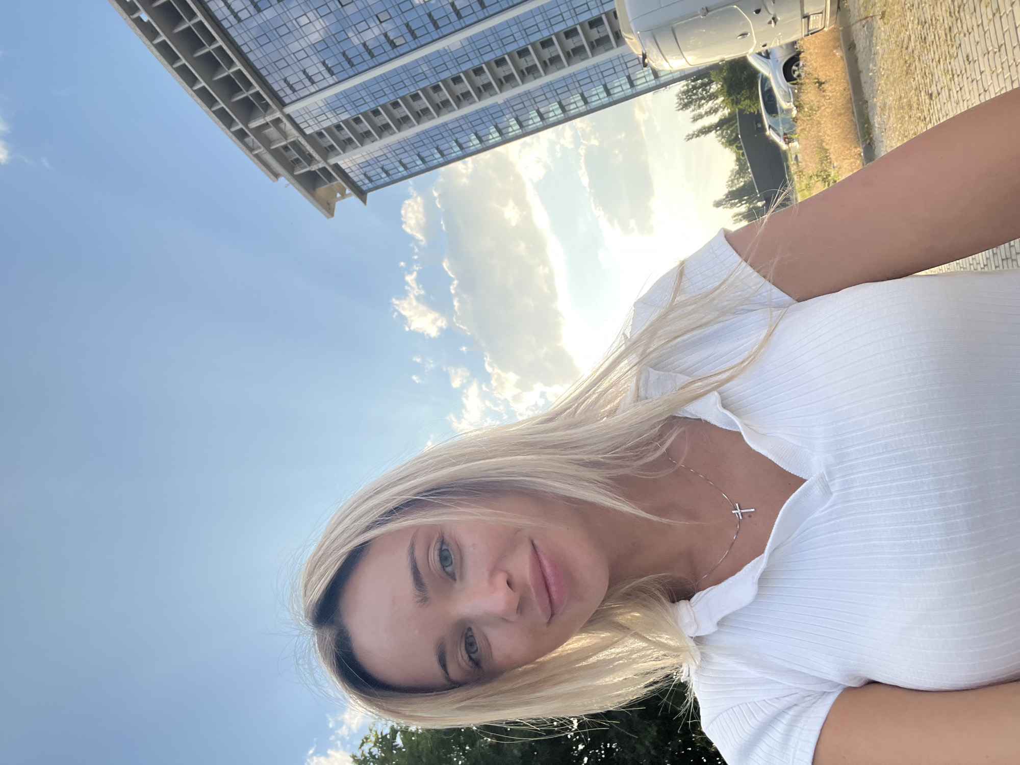In a selfie taken outdoors, a young woman with blonde hair and green eyes smiles at the camera. She is wearing a white, short-sleeved ribbed shirt and a silver cross necklace. The background shows a clear blue sky with scattered white clouds, a tall, unfinished building under construction, trees, and cars parked on a paved surface. The image appears to have been taken during the day, with sunlight illuminating the scene. The location is identified as Fontanka, Ukraine.