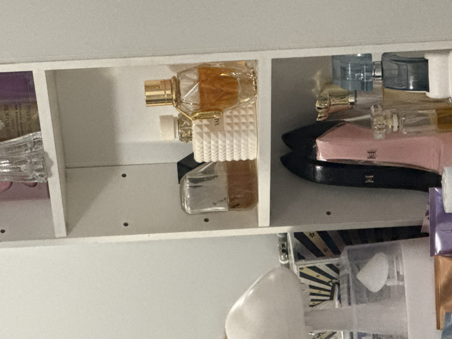 A white cabinet with multiple compartments holds various items, primarily perfume bottles. There are glass bottles with gold accents, a white studded bottle with a black base, and a pink bottle with a black label. Other items include a blue bottle, a clear plastic container with white contents, and decorative boxes with stripes and gold stars. The cabinet appears to be part of a vanity or storage area.