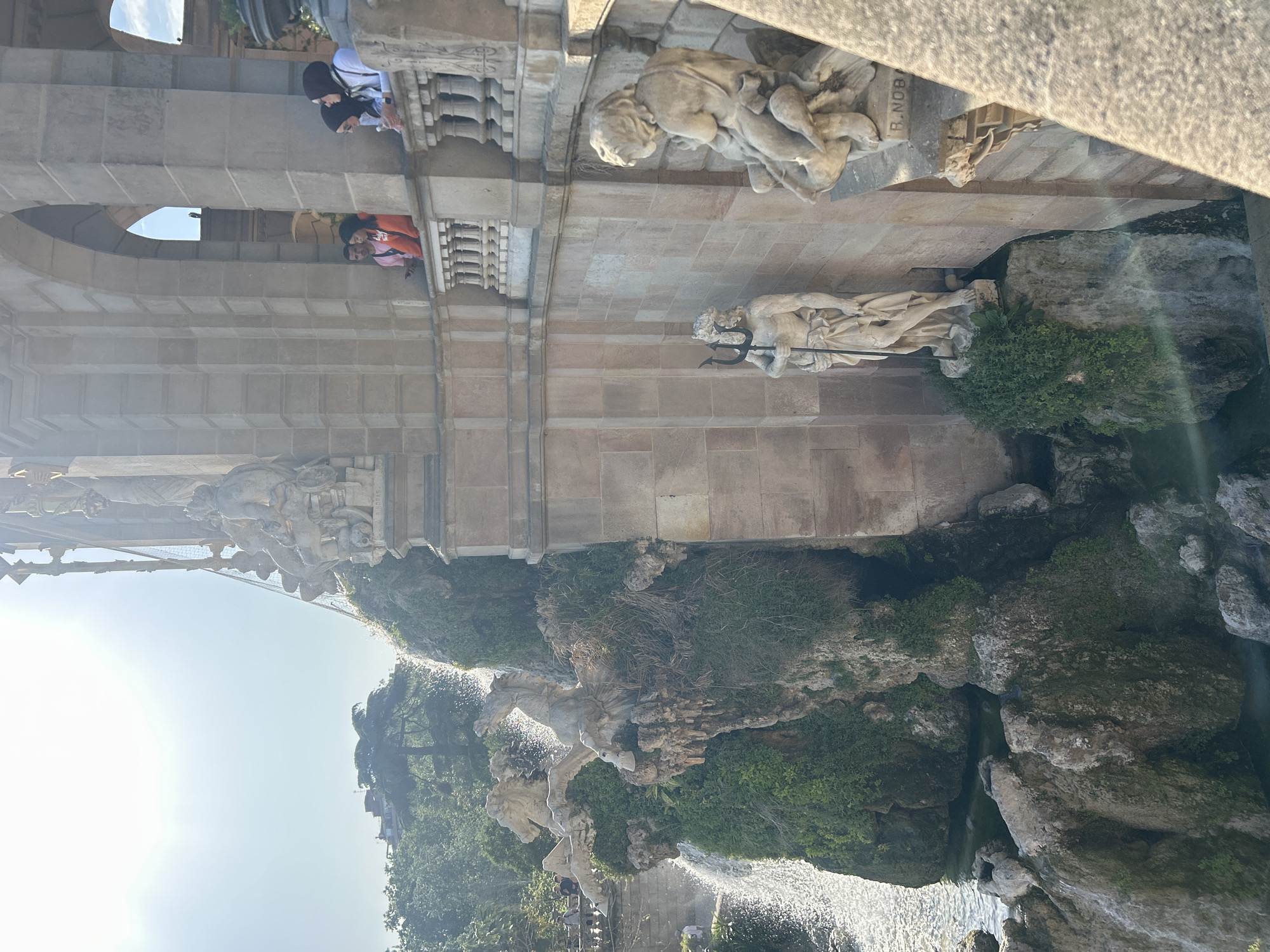 This image shows the Cascada del Parc de la Ciutadella in Barcelona, Spain. The photo is taken from a lower vantage point, looking up at the structure and its various sculptures. Several figures are visible on the upper level of the structure, including people looking over the railing. Below them, sculptures of mythological figures and animals are mounted to the structure, next to cascading water and rock formations covered in green vegetation. Sunlight shines through the scene.