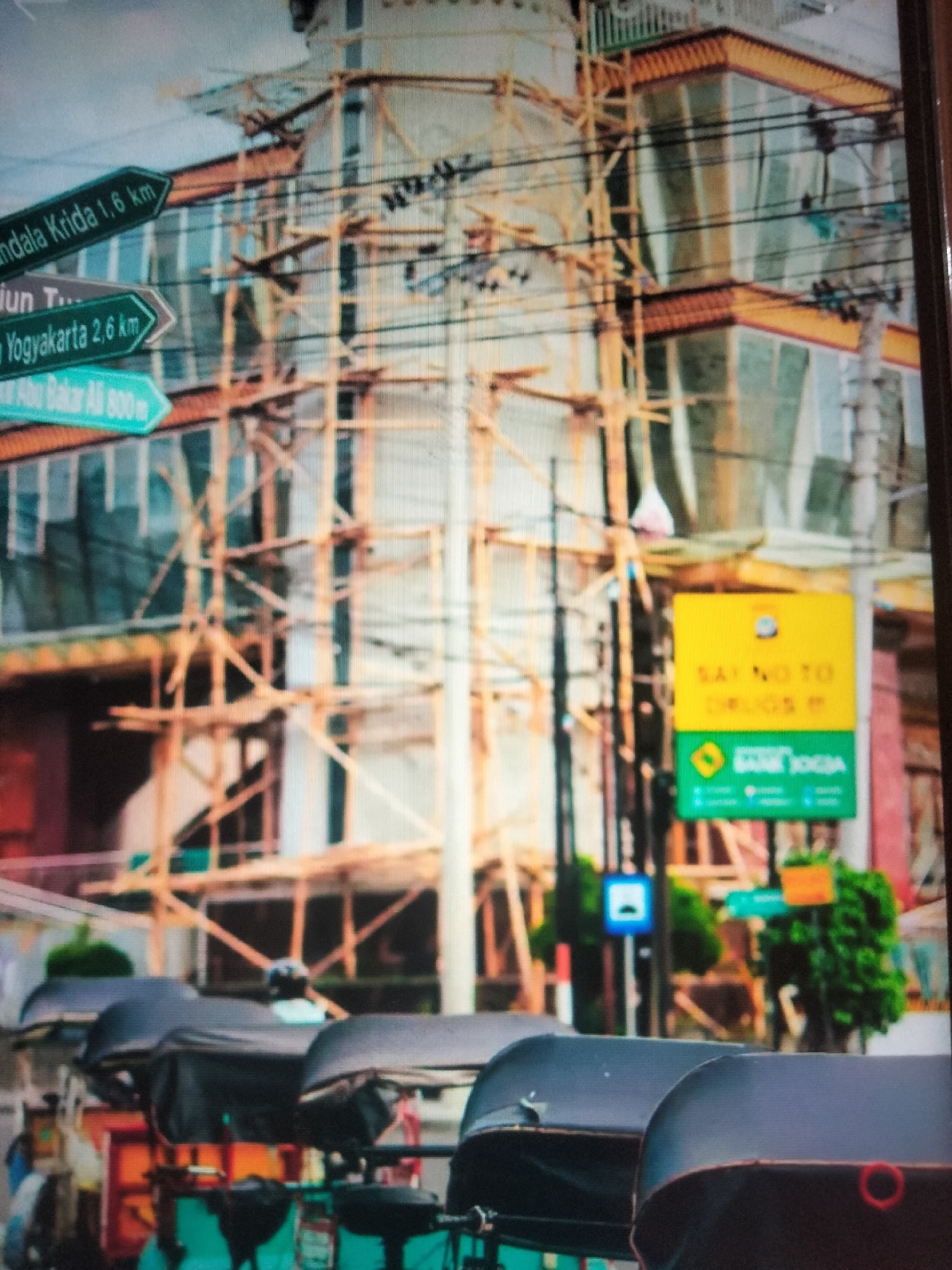 The image depicts a street scene with a building under construction, covered in scaffolding. Several directional signs are visible on the left, indicating distances to locations such as "Mandala Krida 1,6 km", "Yogyakarta 2,6 km", and "Bakar Ali 800m". A yellow and green sign with text that appears to be "SAY NO TO DRUGS" and a bank logo is also present. In the foreground, three parked auto rickshaws with black covers are lined up. The environment suggests an urban setting in Indonesia. The image is pixelated and blurry, making precise identification of details challenging.