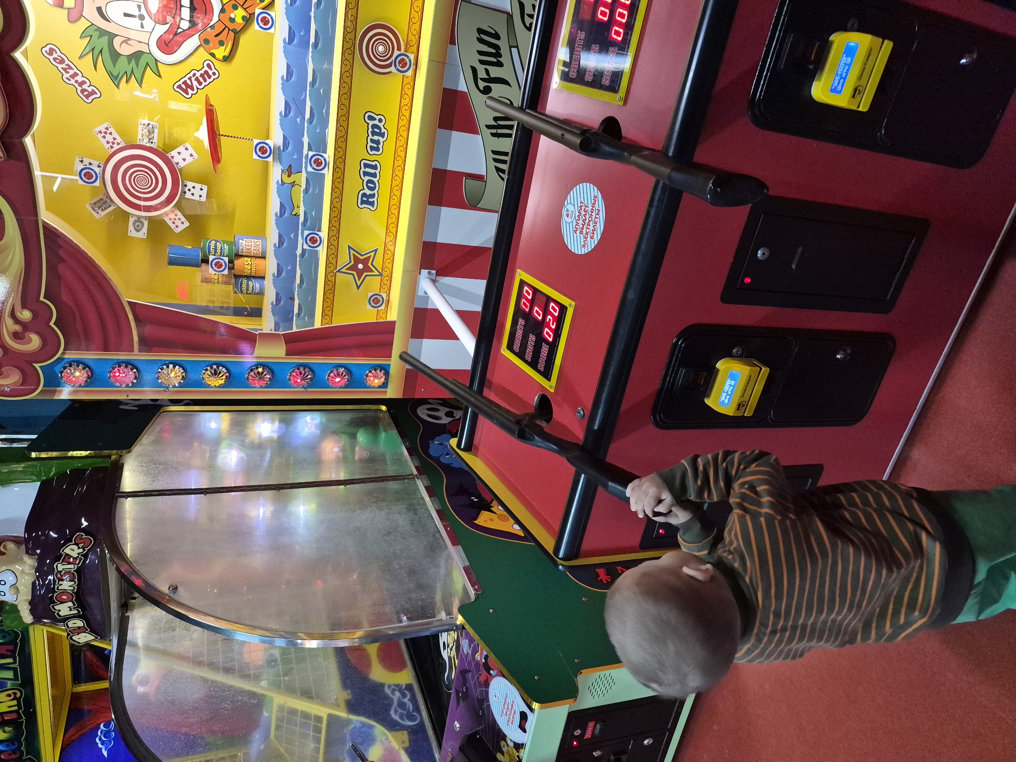 A child in a striped shirt and green pants is playing an arcade game. The arcade game is red, yellow, green, and black, with lights and displays showing "shots" and "score." The game features cartoonish graphics and a shooting element with black rods. Another arcade game with a curved, transparent cover is adjacent. The scene is likely in an entertainment venue. The signs are in English and Russian.