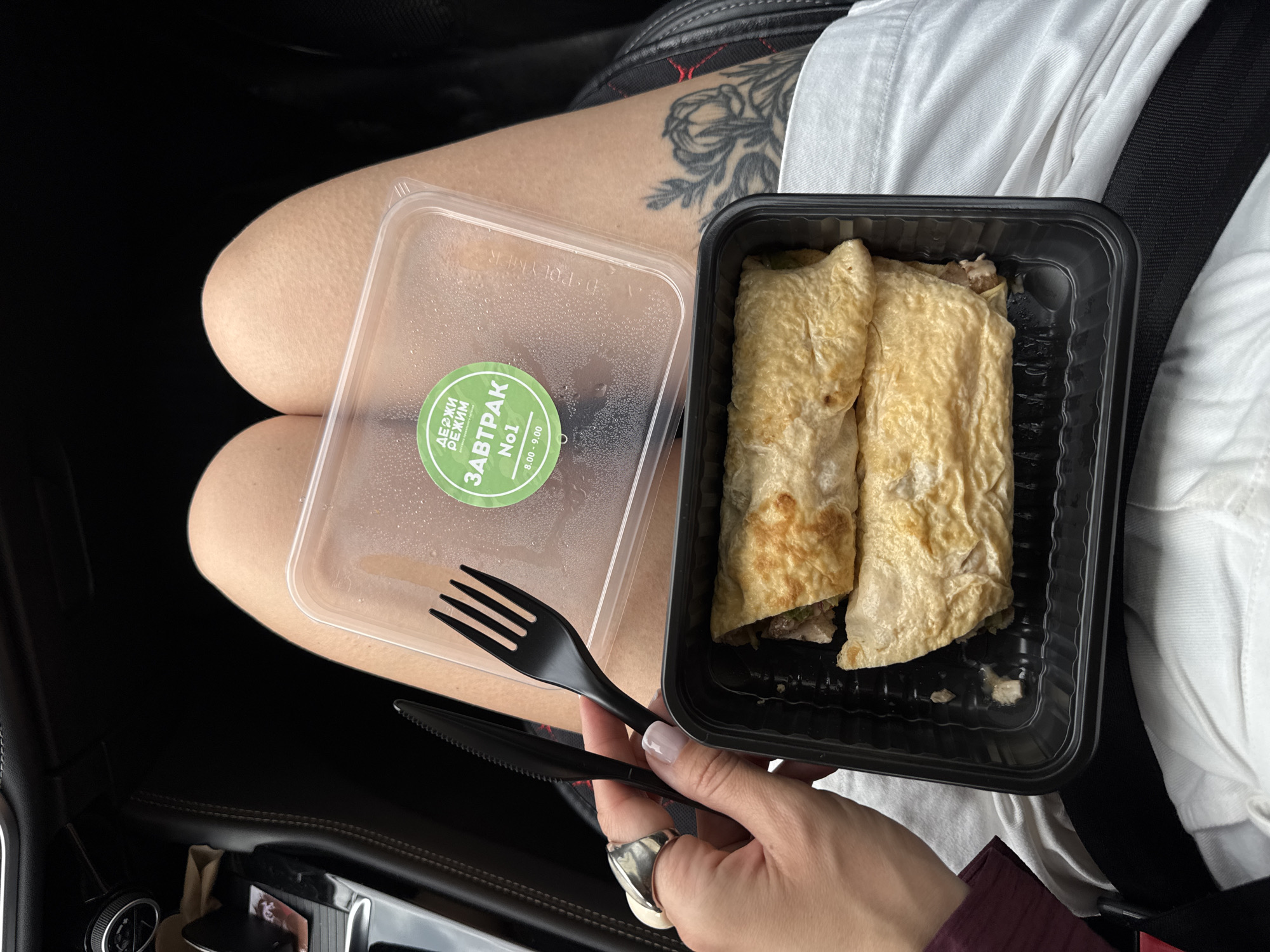 In Sochi, Russia, a person is seated in a vehicle, holding a black plastic fork and knife. In front of them, a black plastic container holds two rolled food items, possibly burritos or wraps, with visible filling. A clear plastic lid, adorned with a green circular sticker bearing text in Cyrillic script, rests beside the food container. The sticker features the words "ДЕРЖИ РЕЖИМ" at the top and "ЗАВТРАК No1" below, indicating "Keep the Regimen" and "Breakfast No1". Below "Breakfast No1" are the numbers "8.00 - 9.00", suggesting a time frame. The person is wearing a large silver ring on their left index finger. A portion of the person's legs is visible, with a floral tattoo on the right thigh. The interior of the vehicle, including dashboard elements and car seats, is also visible.