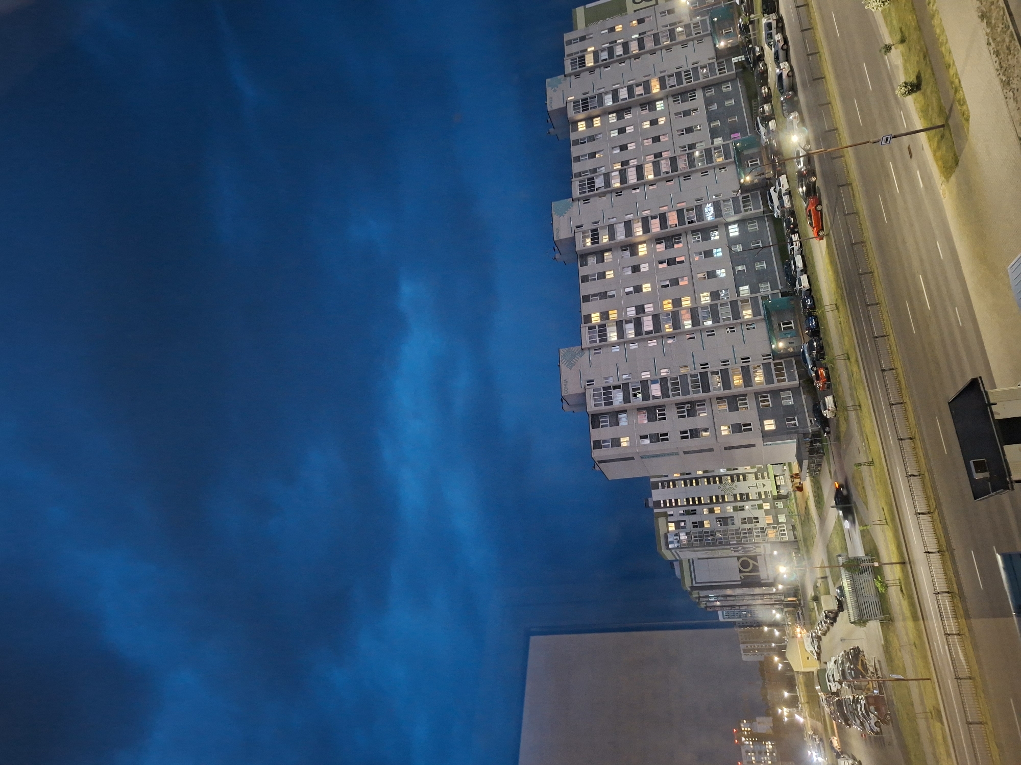 The image depicts an urban scene at night, possibly from a high vantage point. A large apartment building with numerous illuminated windows dominates the right side of the frame. Adjacent to the building is a street with cars parked along the side and driving on the road. Streetlights illuminate the scene. The left side of the frame is filled with a dark blue sky, possibly indicating late evening or early morning. A lower building is visible in the center lower side of the image. The location is in a city, likely within an urban residential district, in Unknown, Unknown.