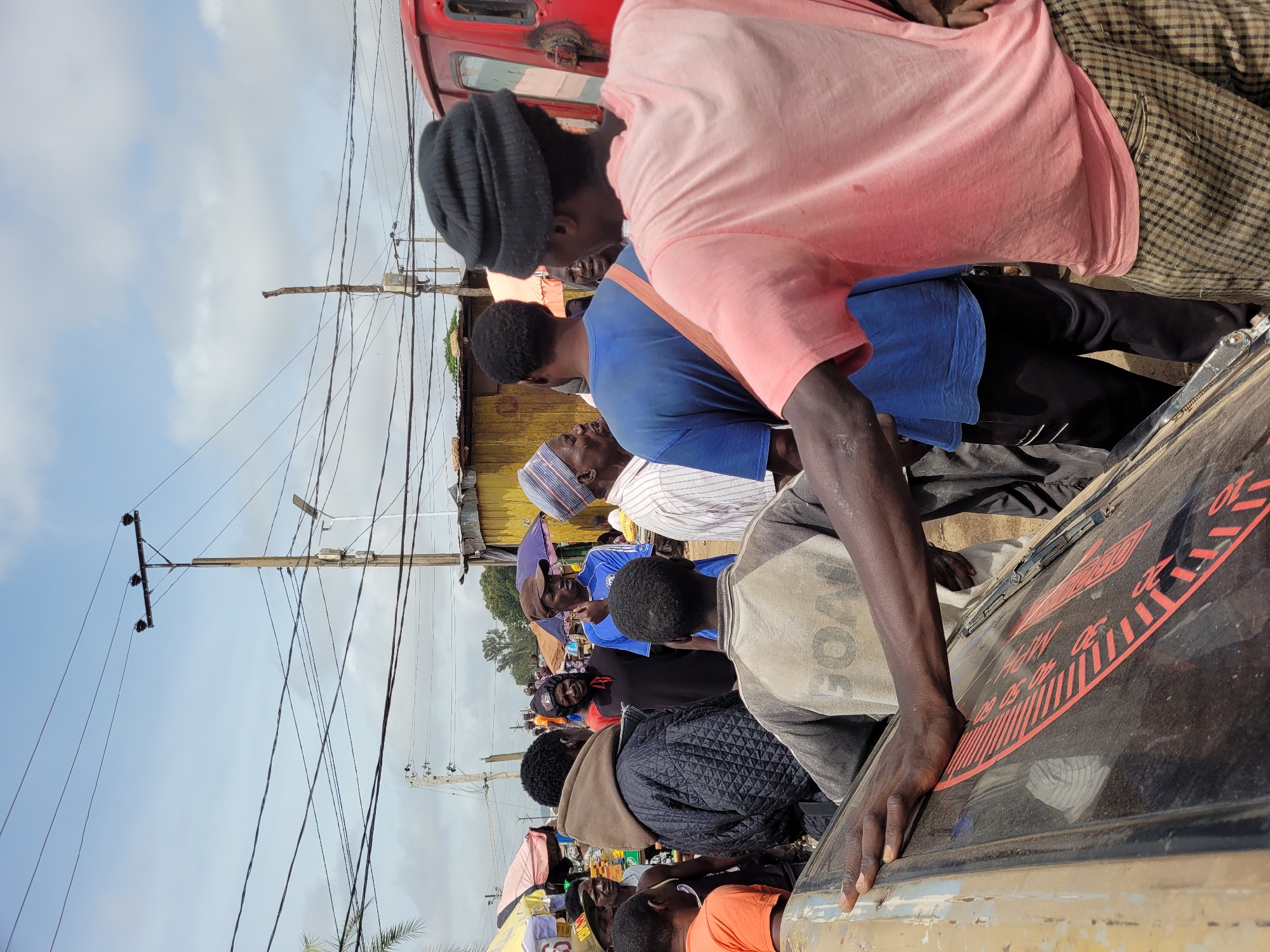 The image depicts a bustling outdoor scene, likely a market or roadside gathering, in Bukuru, Nigeria. A group of individuals are gathered, with most facing towards the center of the composition, suggesting interaction or observation of something unseen.
In the foreground, a person wearing a pink t-shirt and checkered pants is partially visible, leaning on a dusty, tilted vehicle. Their arm is extended, resting on the vehicle's surface, which has a red graphic design. Behind them, another person in a blue t-shirt and black pants is looking to their right.
A man in a quilted black jacket, with a hood pulled up, is prominent in the mid-ground, looking towards the left. He is surrounded by other men. One man wears a blue shirt with a white logo, and another, further back, wears a blue shirt and a darker head covering. A person in an orange shirt is visible on the far left.
The environment includes overhead power lines strung between wooden and metal poles. In the background, makeshift structures with corrugated metal roofs and colorful market umbrellas are visible. The sky is partly cloudy. The overall impression is one of active street life in a densely populated area.