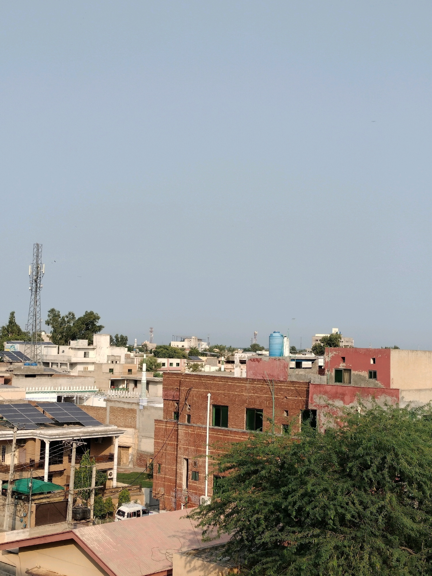 The image depicts a cityscape in Sargodha, Pakistan, under a clear, pale blue sky. The foreground is dominated by a large, bushy green tree on the right. Behind it, several buildings are visible. A prominent red brick building with several windows occupies the center-right. Adjacent to it, to the left, is a building with reddish-pink painted walls and more windows. Further left, a multi-story building with white and light beige walls features solar panels on its roof and a large cellular tower stands tall in the distance on the left side. Several other residential buildings with varying architectural styles and colors are scattered throughout the scene. A white van is parked on a street or driveway in front of a house with solar panels. The overall impression is that of a residential area within a Pakistani city.