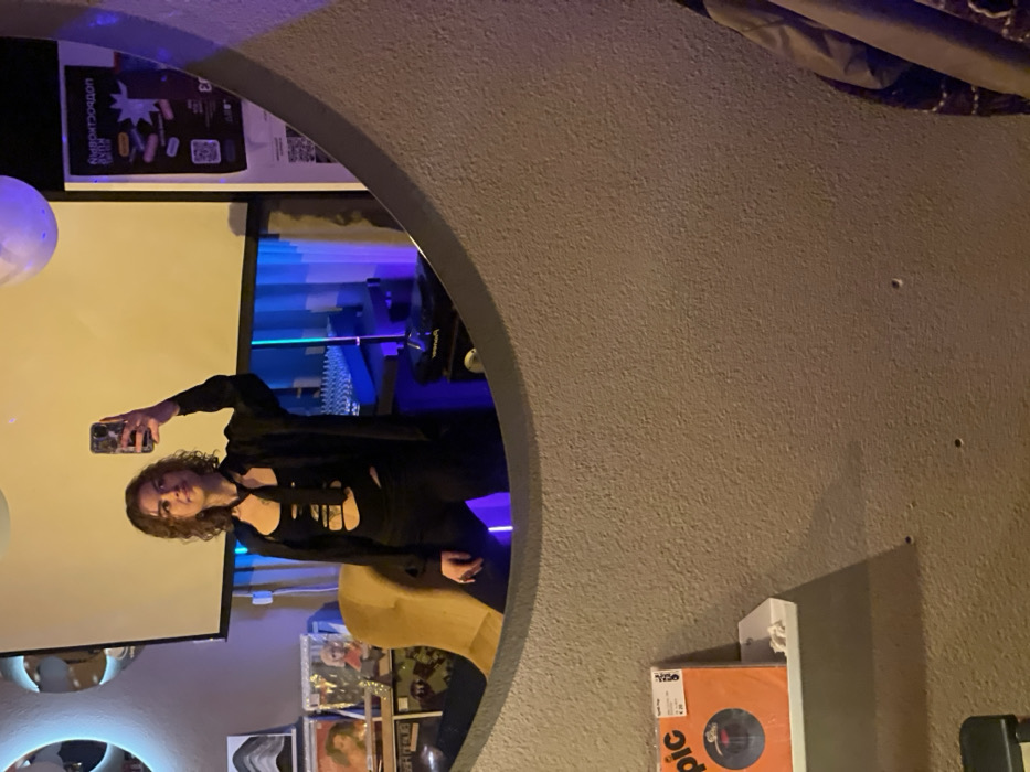 A woman in a black outfit poses for a mirror selfie, holding her phone up. The reflection shows a room with a projector screen, blue lighting, and record albums. A curved gray wall and the edge of the mirror frame dominate the foreground. The overall lighting in the room is dim.