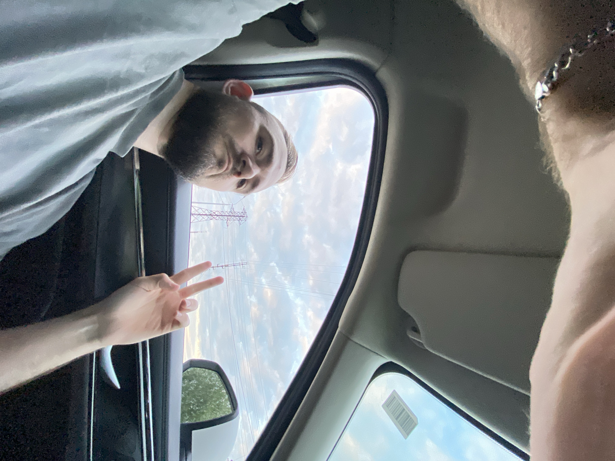 The image depicts a man inside a vehicle, likely a car, with his hand extended out the window, making a peace sign. He has a beard and is wearing a gray t-shirt. The background shows a cloudy sky with power lines and towers, indicating an outdoor, possibly semi-rural or industrial, setting. The interior of the car is visible, including the door handle and a portion of the car's interior lining. The lighting suggests it is daytime. There is no specific indication within the image that places the location in Nyagan, Russia.