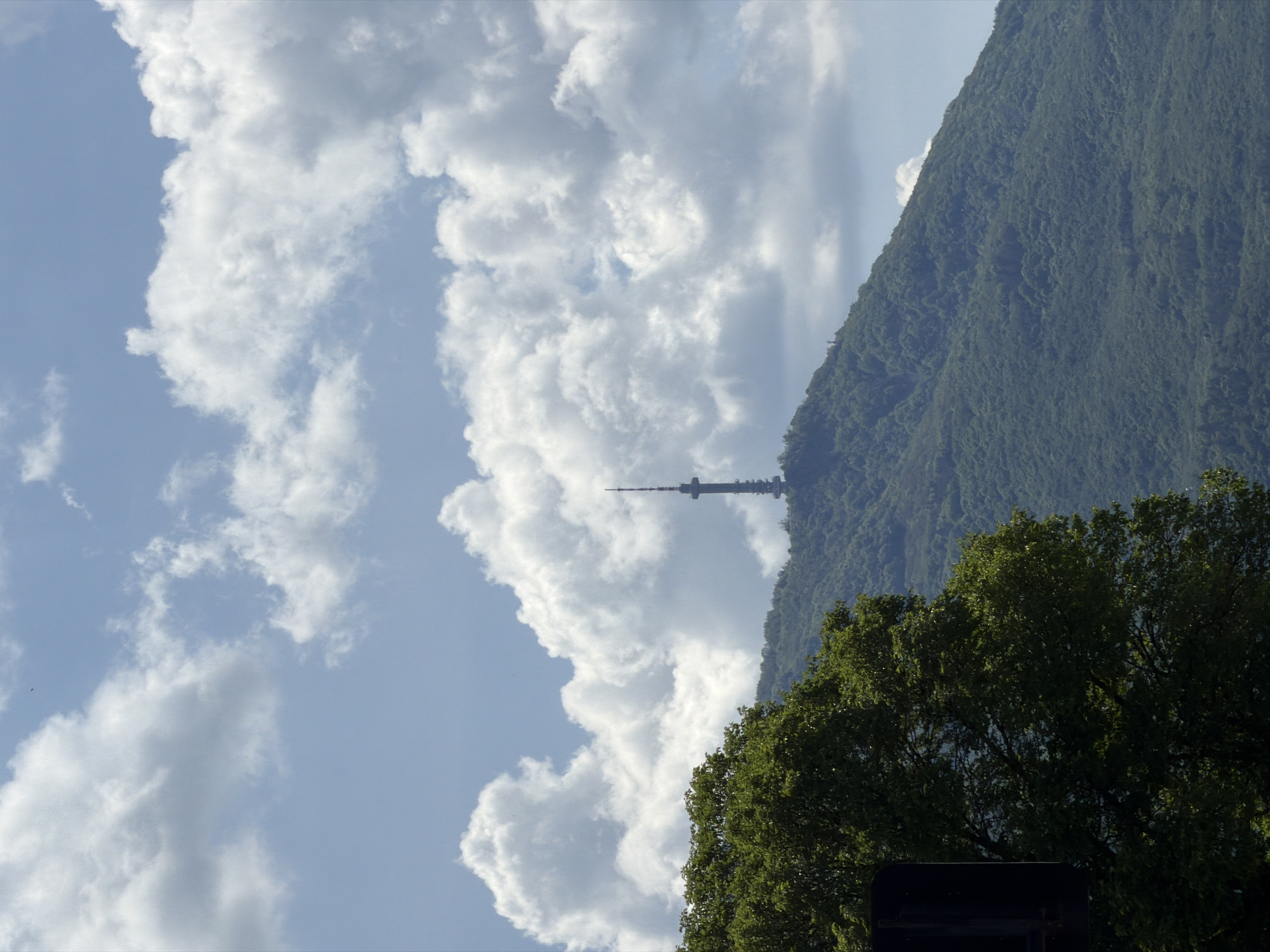 The image captures a daytime scene in Sofia, Bulgaria, featuring the iconic Kopitoto TV Tower perched atop Mount Vitosha. The shot is oriented vertically, showcasing a composition that blends natural and urban elements. Fluffy, white clouds dominate the left side of the frame, contrasting with the clear, pale blue sky. The mountain, covered in dense green vegetation, forms the backdrop on the right. In the foreground, the lush canopy of trees partially obscures the base of the tower. The tower itself stands out as a slender, dark structure against the backdrop of the mountain, signifying the city's telecommunications infrastructure.