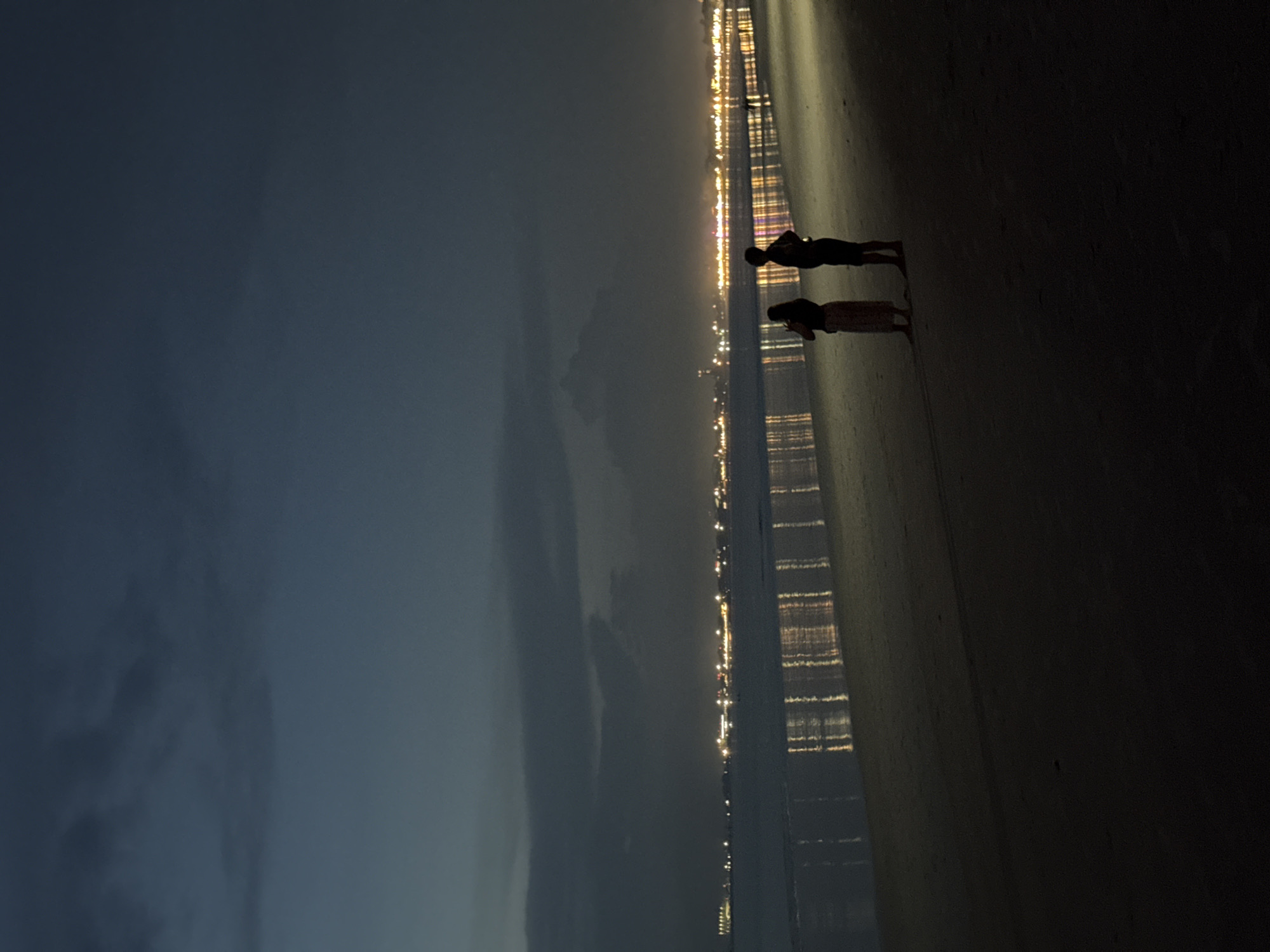 The image shows a dark scene at Kuta Selatan, Indonesia. A man and a woman stand on a beach with their backs to the camera. They appear to be looking toward a city line with lights. The city lights reflect on the wet beach. The sky is dark blue and cloudy.
