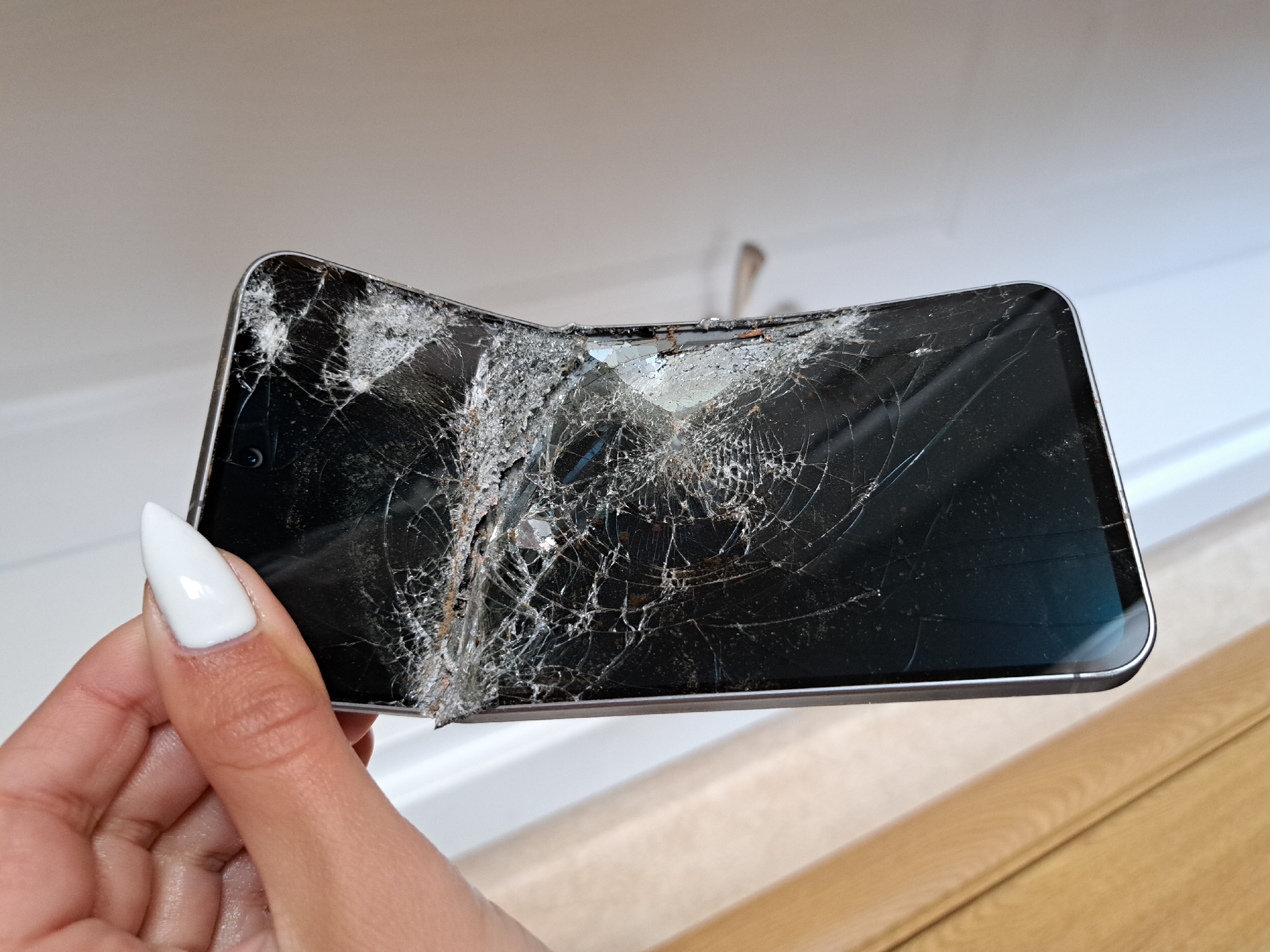 A hand with a white fingernail is holding a severely damaged black smartphone. The screen is shattered and bent, with numerous cracks and displaced glass shards. The phone's frame appears silver. The background is out of focus, showing a white door frame and a wooden surface.