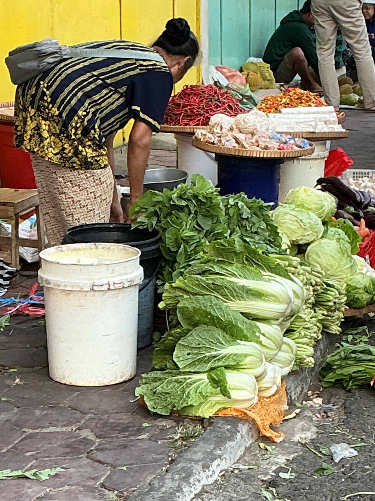 A woman wearing a patterned shirt and skirt with a gray bag slung over her shoulder is bent over a table filled with vegetables at an outdoor market. In the foreground, a white bucket sits on a cobblestone ground, next to a black bucket. Piles of green leafy vegetables, including what appears to be napa cabbage and other greens, are arranged on the ground and on tables. Behind the woman, a man in a green hoodie and light-colored pants is partially visible, sitting on the ground. Other produce, including durian and chili peppers, are displayed on tables in the background. The market appears to be located on a street with a yellow building on the left and a turquoise building in the background. The overall scene depicts a bustling market in Magelang, Indonesia.