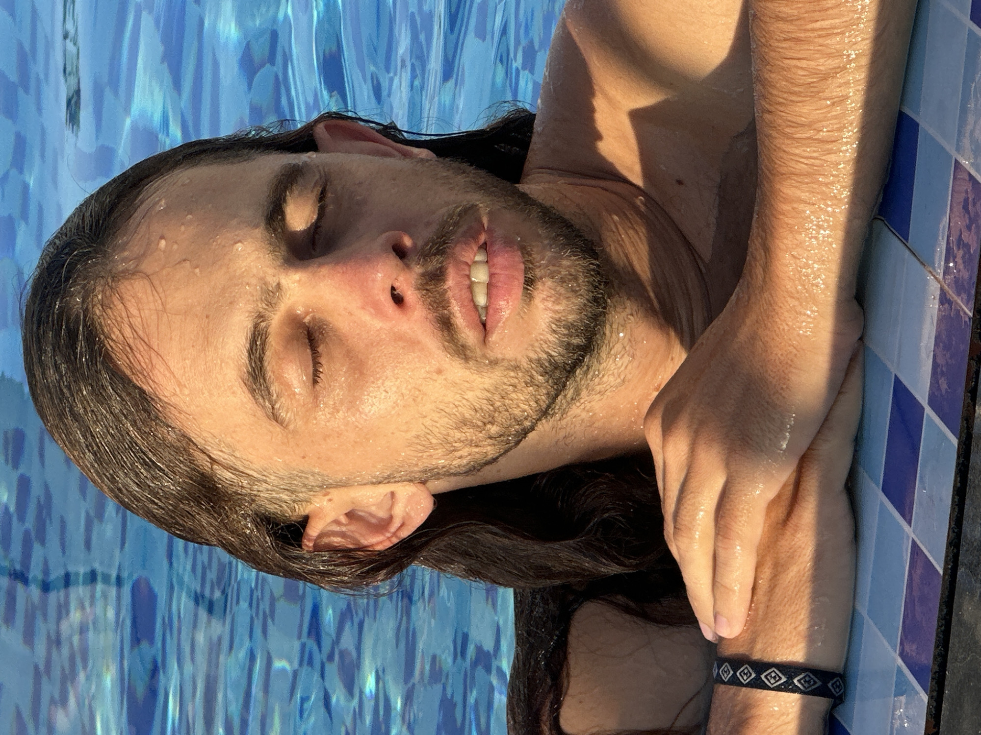 A man with long, dark, wet hair and a beard is pictured from the chest up, submerged in a swimming pool. His eyes are closed, and he appears relaxed. Water droplets are visible on his skin and hair. He is leaning on the edge of the pool, which is tiled with blue and white squares. A dark bracelet with a diamond pattern is on his left wrist. The water in the pool is a bright blue. The lighting suggests it is daytime, possibly late afternoon due to the shadows on his chest. The image does not contain any elements that would allow for identification of a specific location within Đà Nẵng, Vietnam.