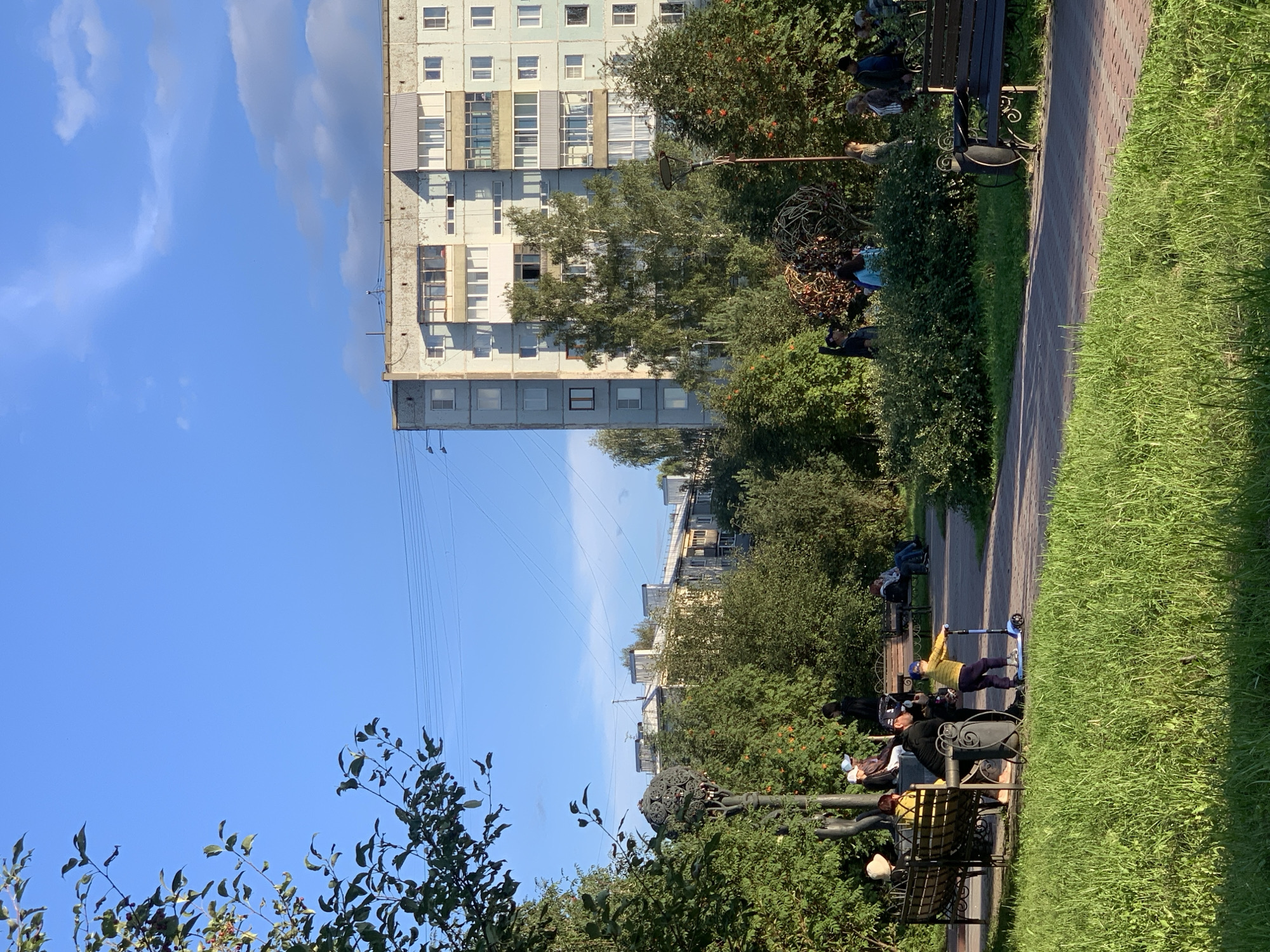 The image captures a sunny day in a park-like setting with a residential building in the background. In the foreground, a wide patch of lush green grass is visible, with a paved walkway winding through it. Several people are enjoying the outdoors on benches and near a lamppost. On the left side of the walkway, a group of people are seated on a bench. Further along, a child is riding a blue scooter, accompanied by an adult. The middle ground is filled with trees and bushes, some bearing red berries. In the background, a tall, multi-story apartment building with a weathered facade stands against a clear blue sky with scattered clouds. The building features multiple balconies and windows. The overall scene suggests a typical urban environment in Prokopyevsk, Russia, with residents enjoying a pleasant day in a public space.