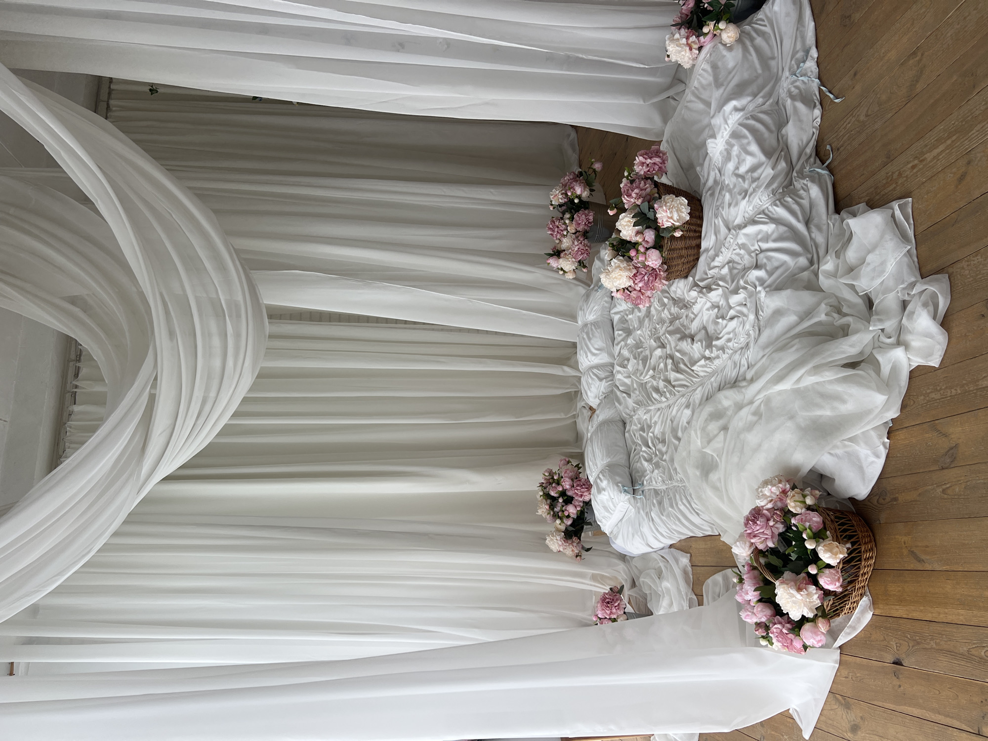 This image depicts a bedroom scene set against a backdrop of flowing white curtains, suggesting a stylized or staged environment. The focal point is a bed covered in white linens, which appear rumpled and draped, creating a soft, luxurious texture. The bed is adorned with several pillows, one of which has light blue ribbons. Scattered around the bed are arrangements of pink and white peonies in woven baskets and a metal bucket. The floor is made of polished wooden planks. The setting evokes a sense of romance and tranquility. There are no people visible in the image, and no specific urban context is apparent, so it is not possible to determine the location as Kyiv, Ukraine.